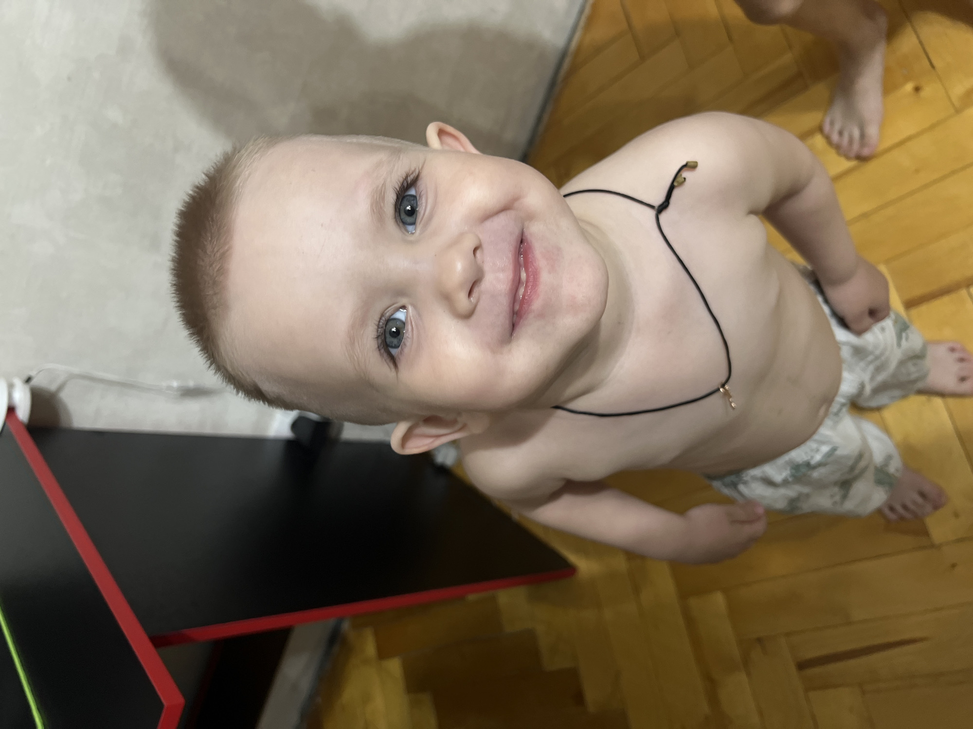 A young child, likely a toddler, smiles upwards at the camera. The child has blonde hair, fair skin, and blue eyes. The child wears a thin black necklace with a small charm. The child is dressed in light-colored camouflage shorts and is shirtless. The child appears to be indoors, standing on a wooden floor. In the periphery of the shot, a black desk with red trim is visible. An adult's foot is visible in the background. There is no indication of the city of Марковский, Russia, within the image.
