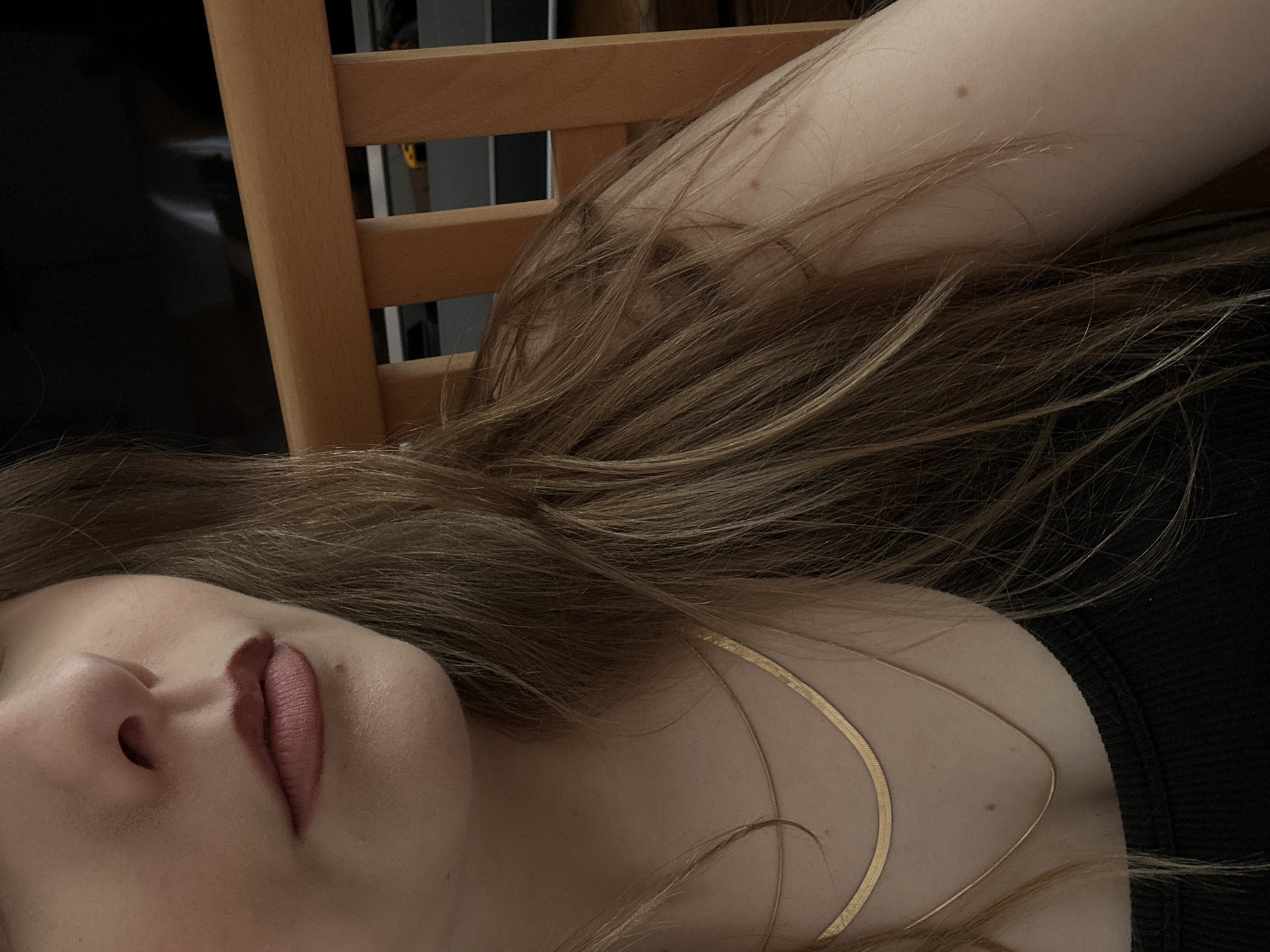 A close-up, low-angle shot shows a woman's face and part of her body. Only her nose and lips are in full focus. She has fair skin and light brown hair, with some strands covering her face and chest. A thin gold necklace is visible on her chest. She is wearing a black top. The background includes a wooden chair and a blurred dark object. No location information about Truchtersheim, France can be determined from the provided image.