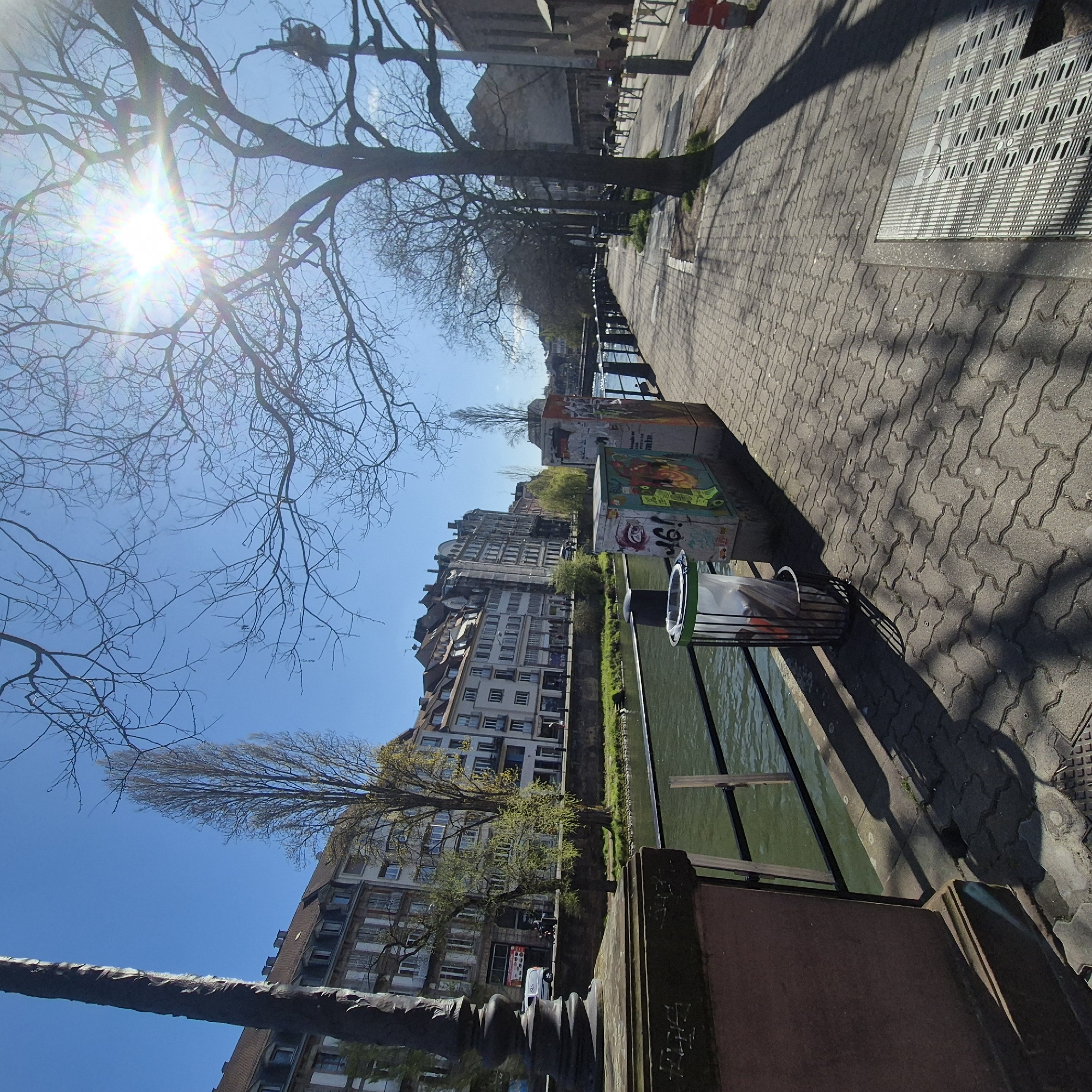 The image shows a view along a canal in Strasbourg, France, characterized by its architecture and waterfront setting. The buildings feature light-colored facades with multiple windows, typical of European urban architecture. Along the canal's edge, there's a paved walkway with patterned paving stones, next to a metal railing. A waste bin is situated near the canal, alongside concrete structures. Bare trees and branches are visible against a clear blue sky, with the sun brightly illuminating the scene. Shadows from the trees are cast on the pavement.