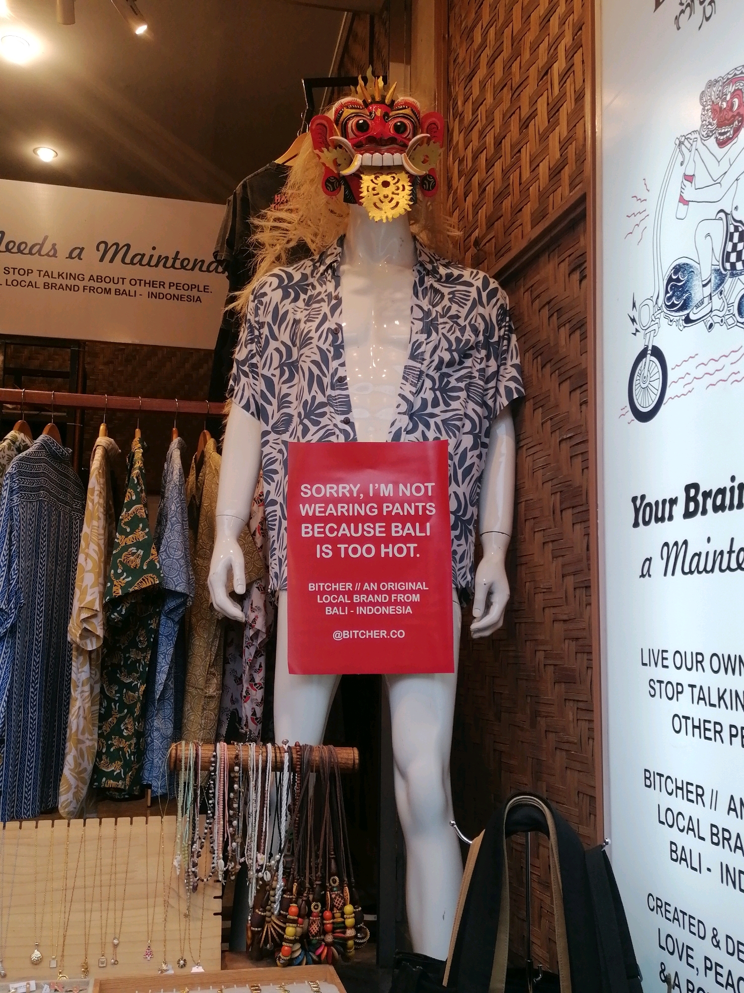 The image shows a retail display in what appears to be a shop in Ubud District, Indonesia. A mannequin is dressed in a patterned short-sleeved shirt and has a mask on its head. A red sign is attached to the mannequin's torso. Behind the mannequin are racks of clothing and a wall with a woven texture. To the right of the mannequin is a large white poster with text and an illustration of a person on a scooter. In the foreground, a display of necklaces is visible. The overall impression is of a fashion or souvenir shop.