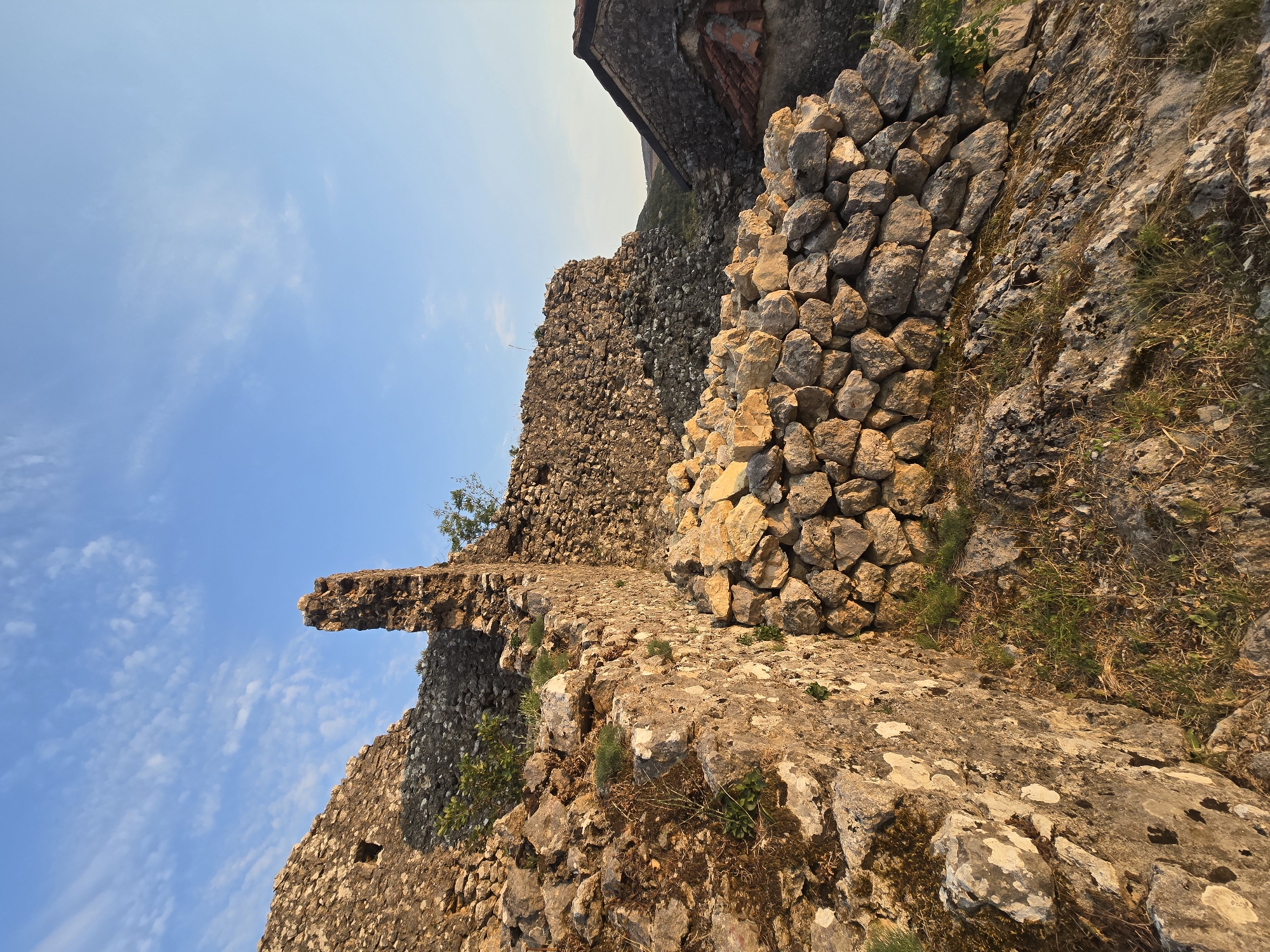 The image depicts the ruins of a stone structure in Medun, Montenegro. The ruins are made of roughly hewn stones of varying sizes, with some moss and vegetation growing on them. In the foreground, a wall of stacked stones forms a barrier. In the background, more crumbling stone walls suggest a larger, possibly fortified, structure. A section of a tiled roof is visible to the right, indicating that some parts of the site may have been more recently inhabited or maintained. The sky above is a clear blue with scattered wispy clouds. The lighting suggests it is either early morning or late afternoon, with warm sunlight illuminating the stonework. The overall scene conveys a sense of historical decay and a rugged, natural environment.