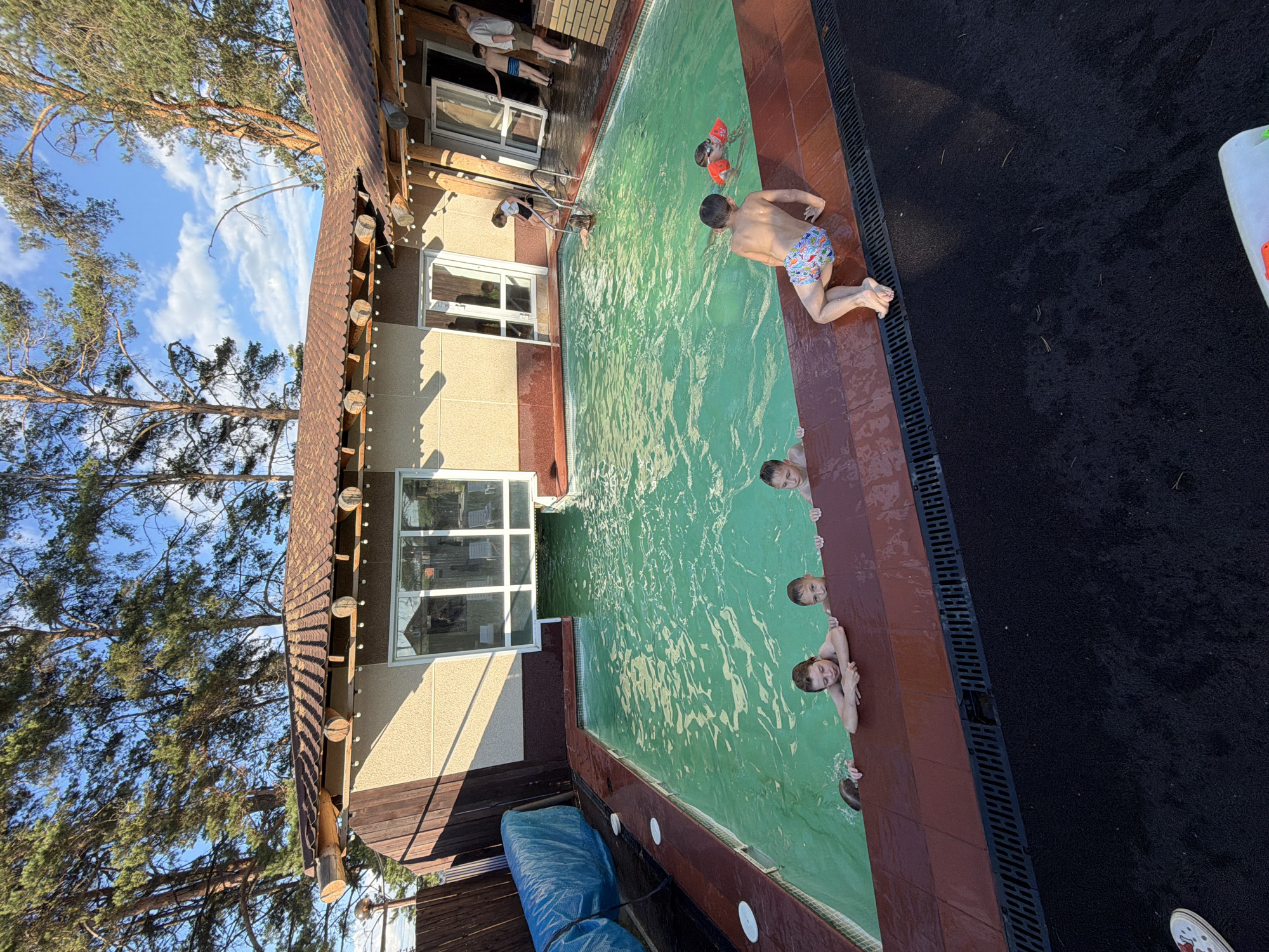 The image depicts a backyard swimming pool area with several children swimming. The pool is rectangular with green water and a brick border. Some children are at the edge of the pool, holding onto the side, while others are in the water. One child in the water is wearing an orange floatation device. A building with a brown tiled roof and large windows is situated behind the pool. Trees are visible in the background. The scene appears to be a private residence in a suburban or rural setting. The location in the city of Chaikovsky, Russia is not discernible from the image itself.
