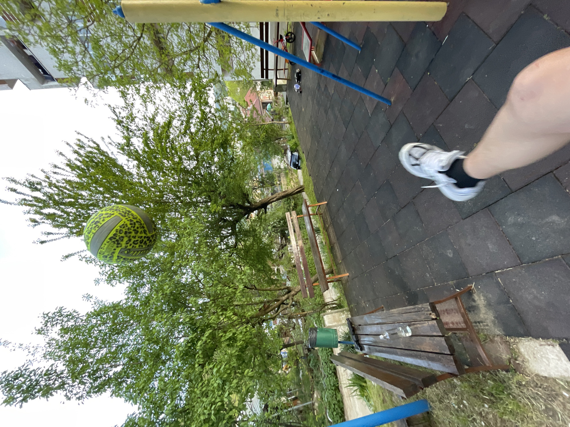 The photograph, taken in Zlatograd, Bulgaria, shows a playground area with interlocking black rubber tiles. A person's leg and foot, clad in a white sneaker with black socks, are visible in the upper right corner of the frame. In the middle ground, there is a green and yellow volleyball with leopard print pattern hanging in front of a tree. The playground equipment, including blue and yellow bars, is visible in the upper portion of the frame. A wooden bench runs horizontally along the bottom edge of the frame. Lush green trees and foliage surround the playground. In the background, buildings and structures suggest a residential area within the city.