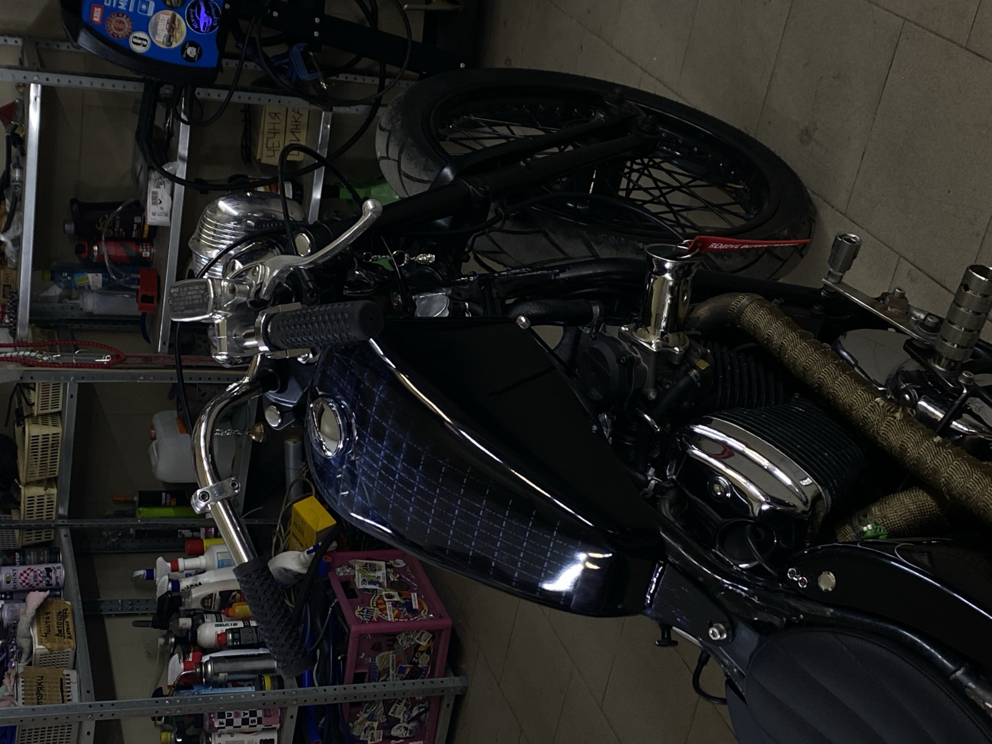 The image depicts a custom black motorcycle parked in a workshop or garage environment. The motorcycle features a custom fuel tank with a dark, patterned design, a chrome engine cover, and a black seat. The front wheel and handlebars are visible, with a small keychain hanging from the left handlebar. Shelving units filled with various items, including spray cans, bottles, and containers, are arranged in the background, suggesting a cluttered workspace. A welding machine or similar industrial equipment is also visible in the background on the right. The floor is tiled. The scene appears to be dimly lit. There are no people visible in the image. The location is identified as Zelenopol'skii, Russia.