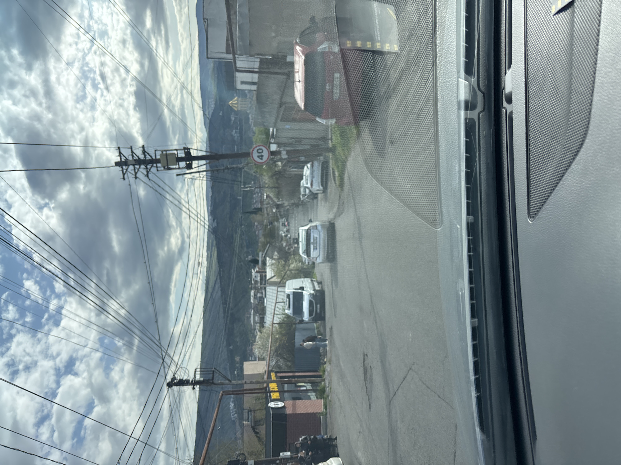 The image, taken from inside a vehicle moving on a road, shows a street scene in what appears to be a residential area. Several parked cars are visible, including a red car and a white van. Power lines are strung across the sky, with a speed limit sign visible beside the road that reads "40". Buildings of various sizes line the street, against a mountain backdrop under a cloudy sky. The location is potentially in or near Tbilisi, Georgia.