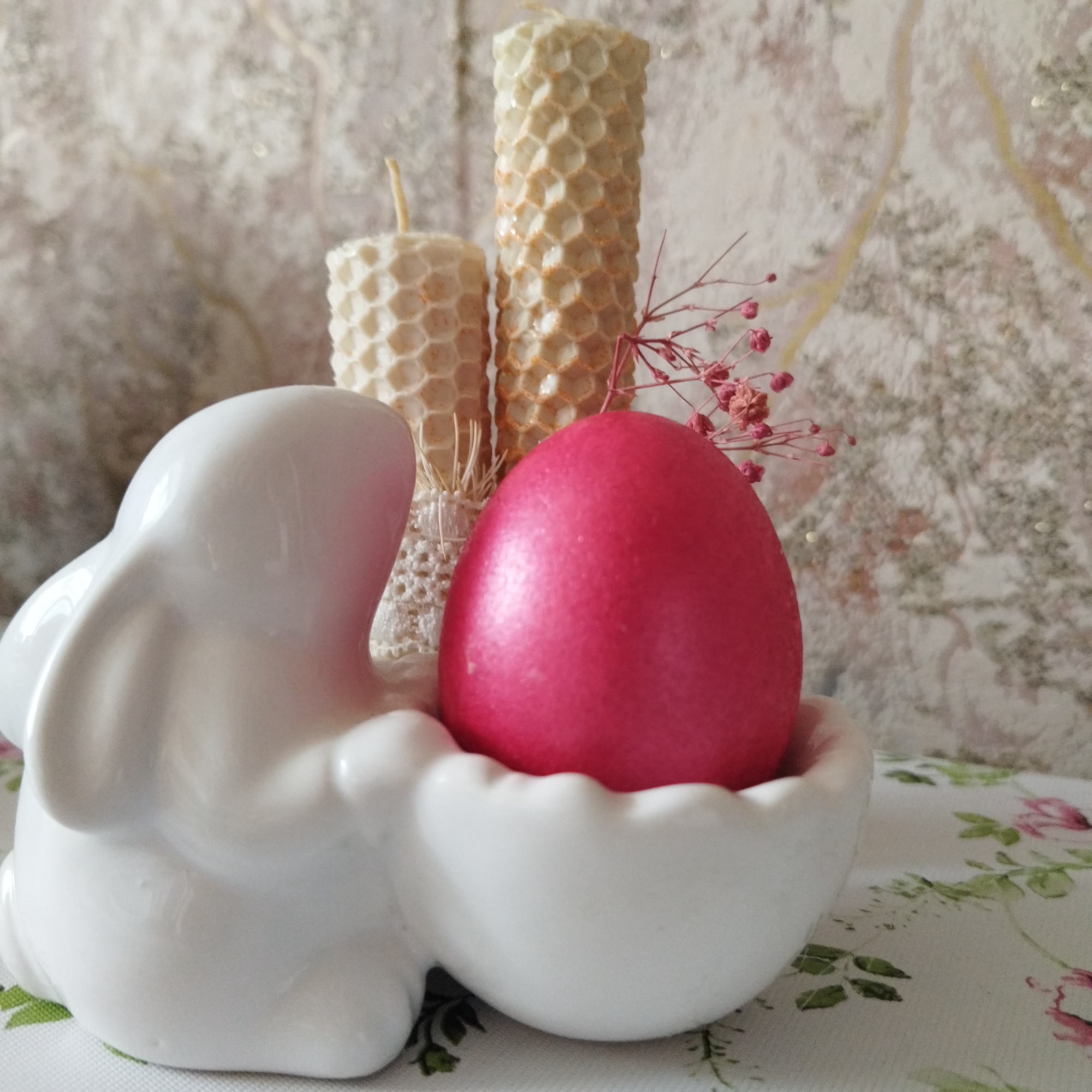 This still-life image displays a festive arrangement, likely for Easter or springtime, set indoors. The primary subjects include a white ceramic bunny figurine, a vibrant pink dyed egg, and two beeswax candles.
The white ceramic bunny, positioned on the left, is designed to appear as if it's holding or cradling the bright pink egg. The egg has a smooth, glossy finish with a subtle shimmer, indicative of a decorated Easter egg. Behind the bunny and egg, two natural-colored beeswax candles stand. Both candles feature a distinct hexagonal honeycomb texture, with one being shorter and wider, and the other taller and more slender. Delicate dried pink floral elements, possibly baby's breath, are subtly placed around the candles and next to the egg, adding a soft pop of complementary color and texture.
The items are arranged on a white surface, which appears to be a tablecloth, adorned with a subtle floral pattern featuring green leaves and hints of pink or red blossoms. The background consists of a light-toned wall or wallpaper with a soft, textured, possibly marbled or abstract design, featuring muted beige or gold undertones. The overall scene evokes a sense of gentle celebration and seasonal decoration. There is no visible text in the image.