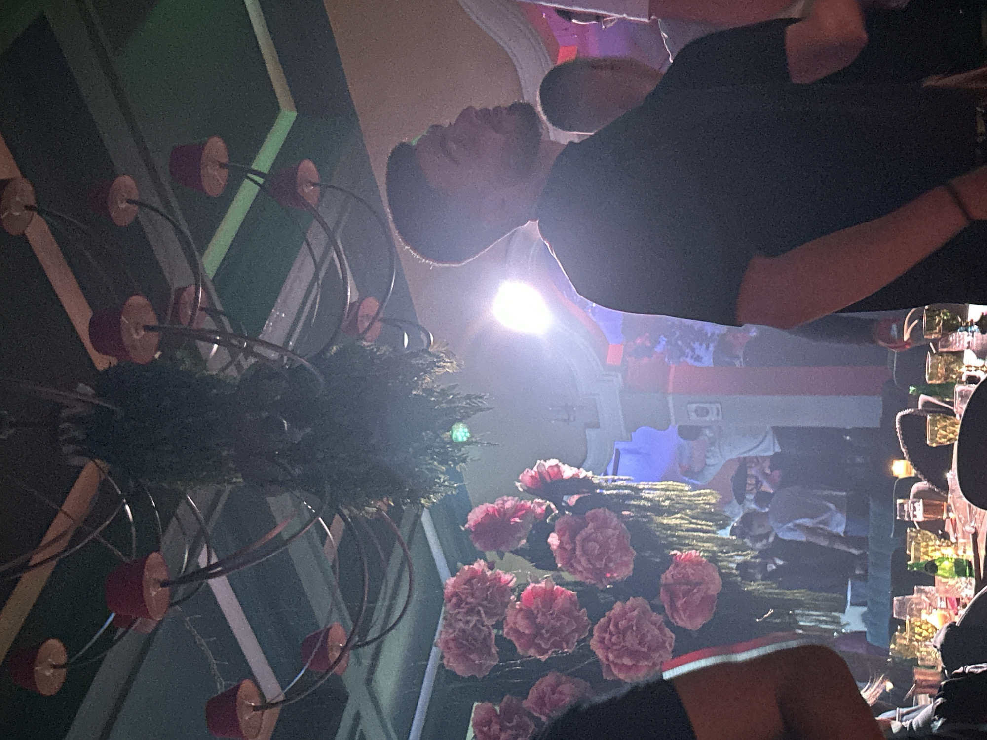 The image shows an indoor scene, possibly a bar or restaurant, based on the presence of a chandelier, flowers, and what appear to be bottles and glasses behind a counter. A man is prominent in the upper part of the frame, wearing a dark shirt. Other people appear in the background. The overall lighting is somewhat dim, with a bright light source creating glare. It is difficult to determine the location as Sofia, Bulgaria with the provided image details.