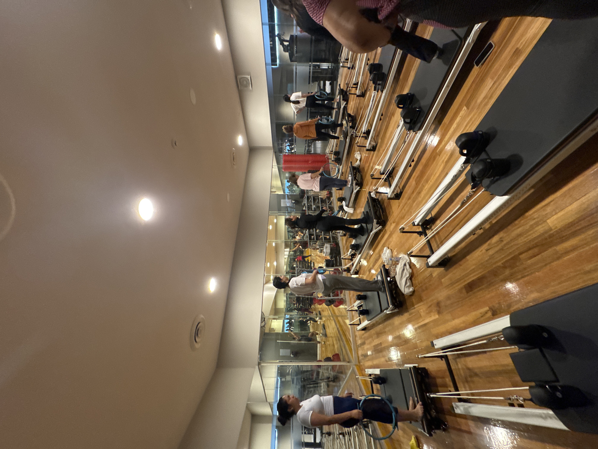 The image shows a pilates studio, likely in a gym or specialized fitness center. Several people are engaged in a pilates class, using reformer machines. The studio features wooden flooring and mirrored walls, and the lighting is bright and artificial. The exact location in the city of Puebla, Mexico cannot be confirmed from the image.