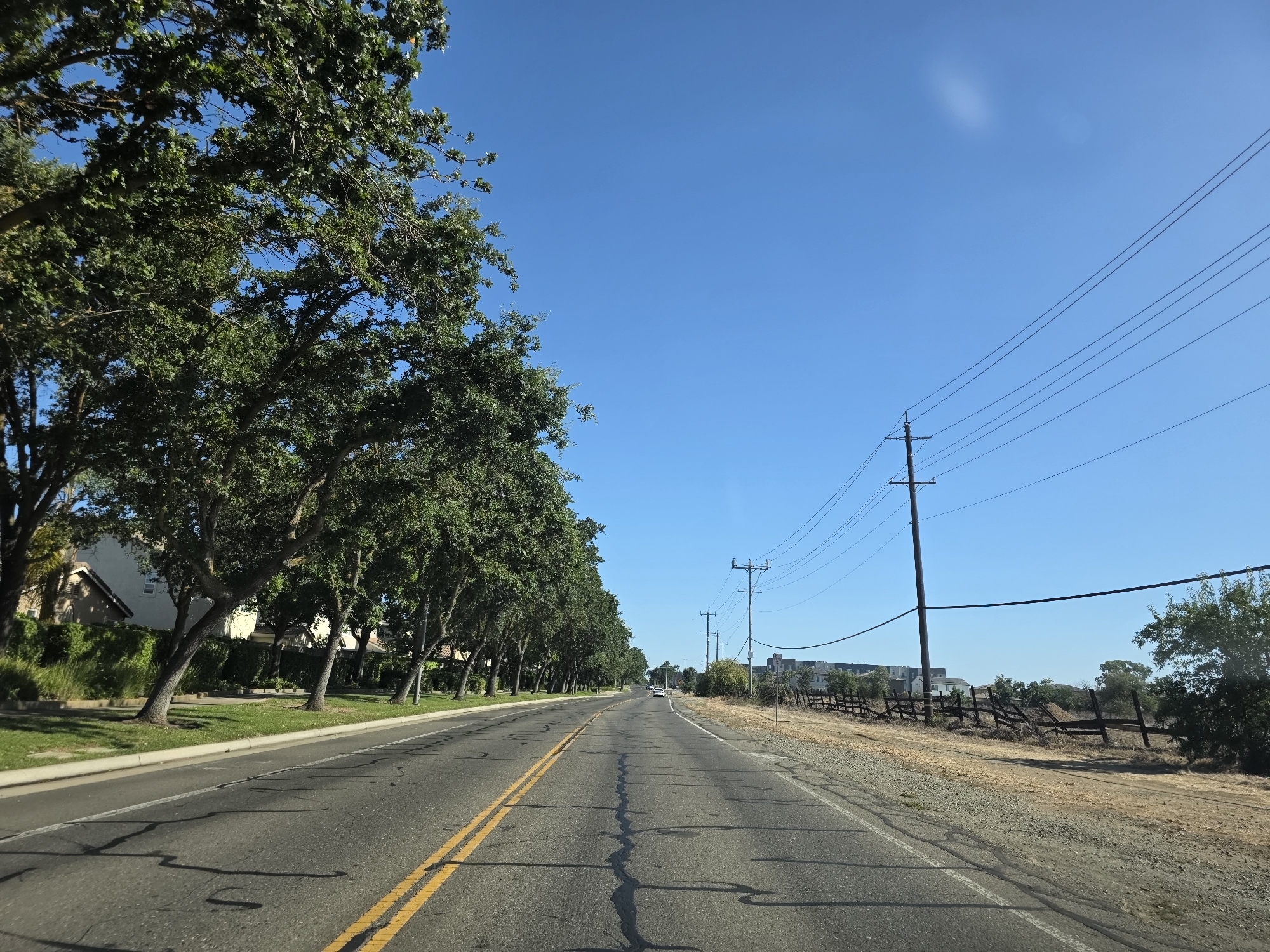 This media file depicts a paved road with a double yellow line in the center, suggesting it is a two-lane road. The road stretches into the distance under a clear, bright blue sky, indicating a sunny day. On the left side of the road, a line of mature trees with lush green foliage forms a canopy, casting dappled shadows on the road. Behind the trees, residential buildings with beige exteriors and pitched roofs are partially visible, hinting at a suburban or residential area.
On the right side of the road, a dry, scrubby embankment is present, with a broken wooden fence running along its edge. Utility poles with power lines are also visible, stretching across the sky and along the right side of the road. In the far distance, a few white cars are seen on the road, and a cluster of modern buildings is visible on the horizon.
The scene appears to be captured from a moving vehicle, as suggested by the perspective and the motion blur of the shadows on the road. The overall impression is one of a pleasant, clear day in a semi-rural or suburban setting, possibly in the United States given the location context. There is no visible text in the image. The time of day is likely late morning or early afternoon, given the bright sunlight and the angle of the shadows. The weather is clearly fair and sunny.