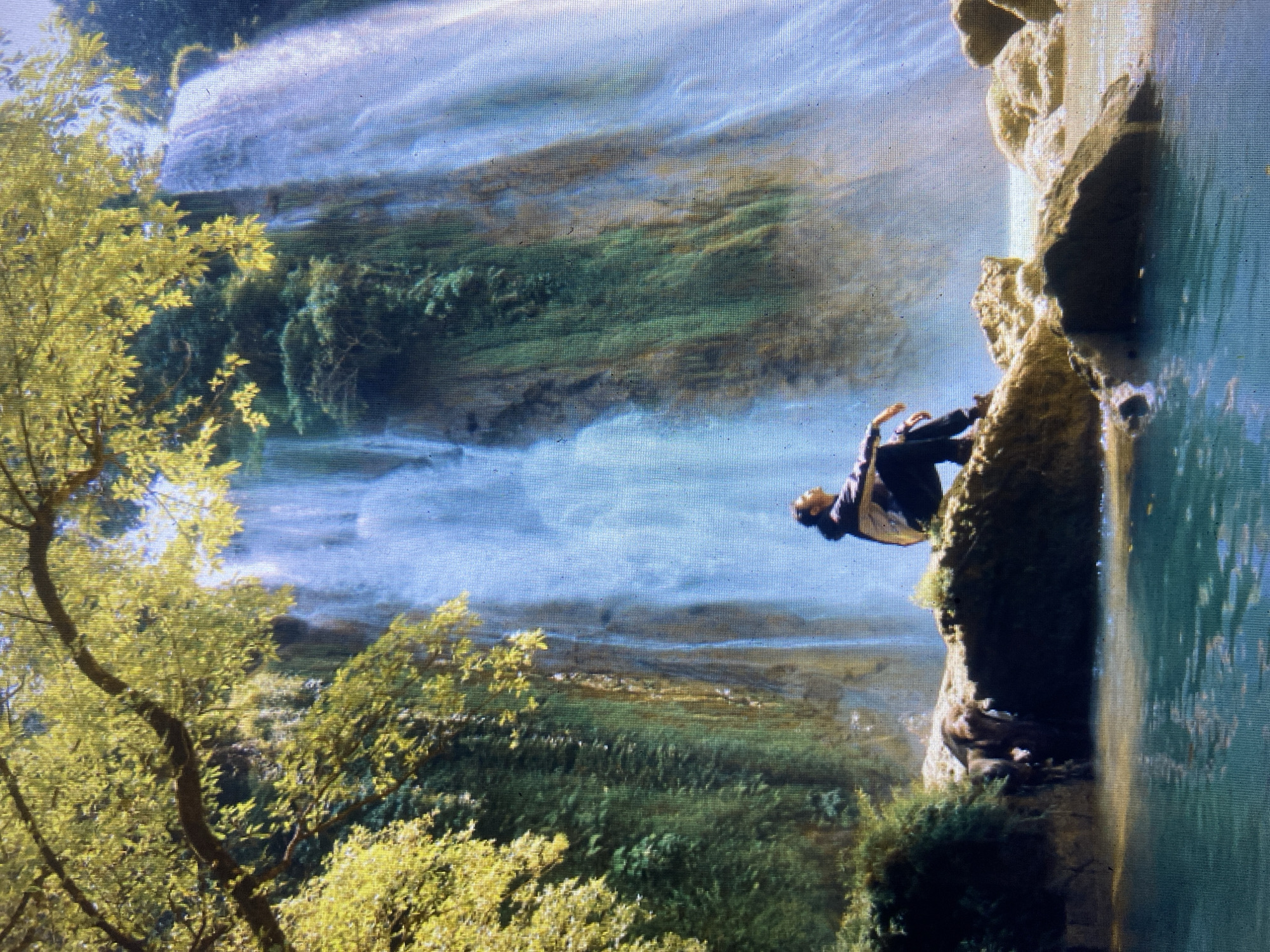In the city of Girijaya, Indonesia, a person is sitting on a large rock near a majestic waterfall. The waterfall cascades down a moss-covered cliff face, creating a misty spray that is illuminated by sunlight. Lush green foliage frames the scene, with a large tree branch extending into the foreground, its leaves catching the light. The water at the base of the waterfall is a vibrant turquoise, reflecting the surrounding greenery and the bright sky. The person, facing away from the camera, is wearing dark pants, a light-colored shirt with a dark hood, and dark shoes. They are sitting with their legs crossed, appearing to be contemplative as they gaze at the powerful display of nature.