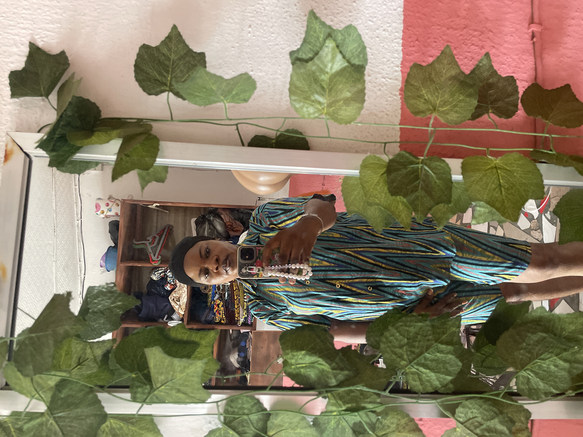 A person is taking a selfie in a mirror. The person is wearing a patterned dress with a black head covering. They are holding a phone with a white beaded strap. Behind them is a shelf with clothes and other items. The mirror is decorated with artificial ivy. The wall behind the mirror is painted white and pink. The location is presumably Badagry, Nigeria.