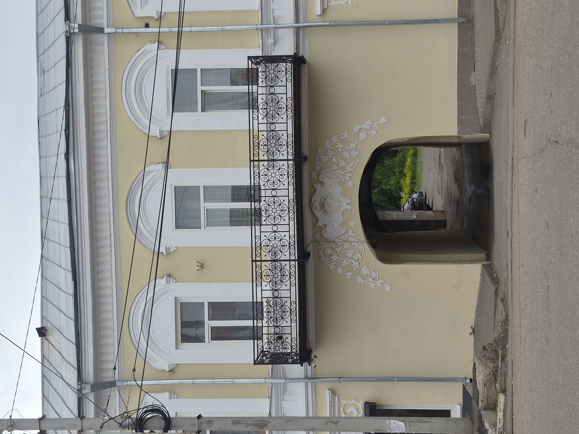 The image depicts a two-story building with a light yellow stucco facade and white architectural trim, situated in Sarapul, Russia, under an overcast sky. The building features a sloped, seamed gray metal roof with a gutter running along its edge. A small, dark rectangular object protrudes from the roof near the left.
The upper floor displays three arched windows with white frames. A black wrought-iron balcony, adorned with circular and scroll-like patterns, extends from below the two rightmost windows. Below the balcony, a large, dark arched passageway pierces the building's center, leading to an interior area. The archway is embellished with ornate white stucco floral and scroll patterns on either side and above its apex. Two gray metal drainpipes are visible; one on the far left, partially obscured by a utility pole, and another to the right of the central facade, descending into the pavement. A small, white, pronged fixture is mounted on the wall between the left and central upper windows.
In the foreground, an asphalt road with visible cracks is present, bordered by concrete curbs and paved sections of sidewalk. A gray concrete utility pole on the left supports numerous black electrical wires and coiled cables. Beyond the archway, a paved drive leads to an area containing several parked dark-colored vehicles, green trees, and yellow flowering plants. No individuals are visible in the scene.