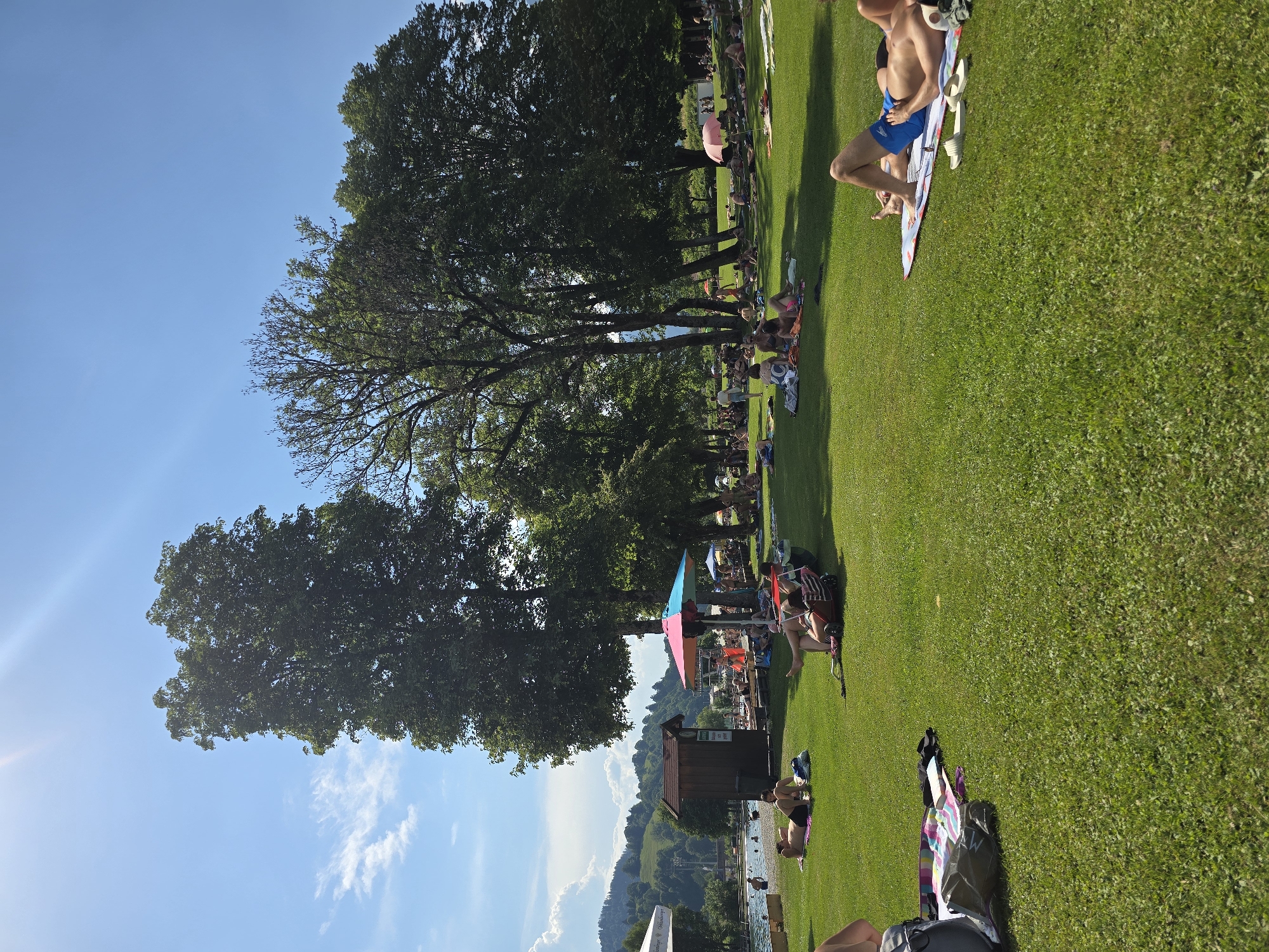 This media file depicts a sunny day at an outdoor recreational area, likely a park or a lakeside beach, situated in Garmisch-Partenkirchen, Germany, given the context. The scene is dominated by a lush green lawn where numerous people are gathered, enjoying the warm weather. The image is oriented vertically.
The main subjects are the people, many of whom are lying on towels or blankets, sunbathing, and relaxing. Some individuals are wearing swimwear, suggesting they are at a swimming area or a place where swimming is permitted. There are also trees providing shade, and umbrellas, including a distinctive multi-colored one, offering respite from the sun. In the background, a body of water can be seen, with more people in and around it. Further back, hills and buildings suggest a town or village nestled in a valley, characteristic of the Bavarian Alps.
The activity taking place is leisure and recreation on a clear, sunny day. The bright sunlight and blue sky indicate fair weather. The presence of a lake or river, the green grass, and the mountainous backdrop strongly suggest a summer day in a scenic, mountainous region. The atmosphere appears relaxed and convivial, with people enjoying their time outdoors.
Notable details include the clear blue sky with a few wispy clouds, the vibrant green of the grass, and the shadows cast by the trees and umbrellas, which help indicate the time of day is likely midday or early afternoon. There are no clearly legible texts visible in the image.