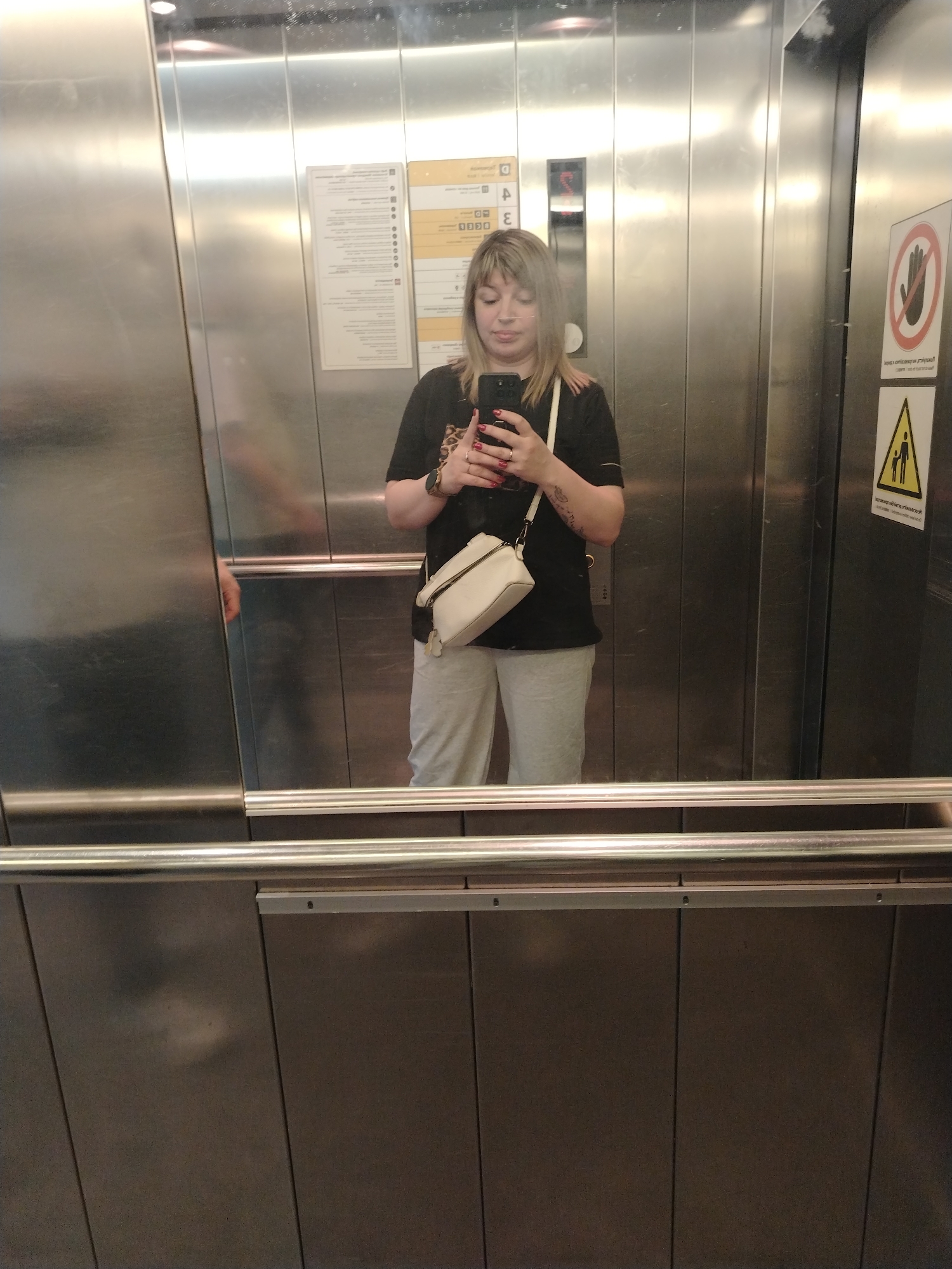 A fair-skinned woman is taking a selfie in an elevator. She has shoulder-length blonde hair with bangs. She is wearing a black t-shirt, light gray sweatpants, and a white crossbody bag. She is holding a smartphone in her hands with red nail polish.
In the background, there are stainless steel elevator walls. On the right elevator wall, there are two warning signs. The elevator's interior appears to be well-lit.
The elevator is located in the city of Khimki, Russia.