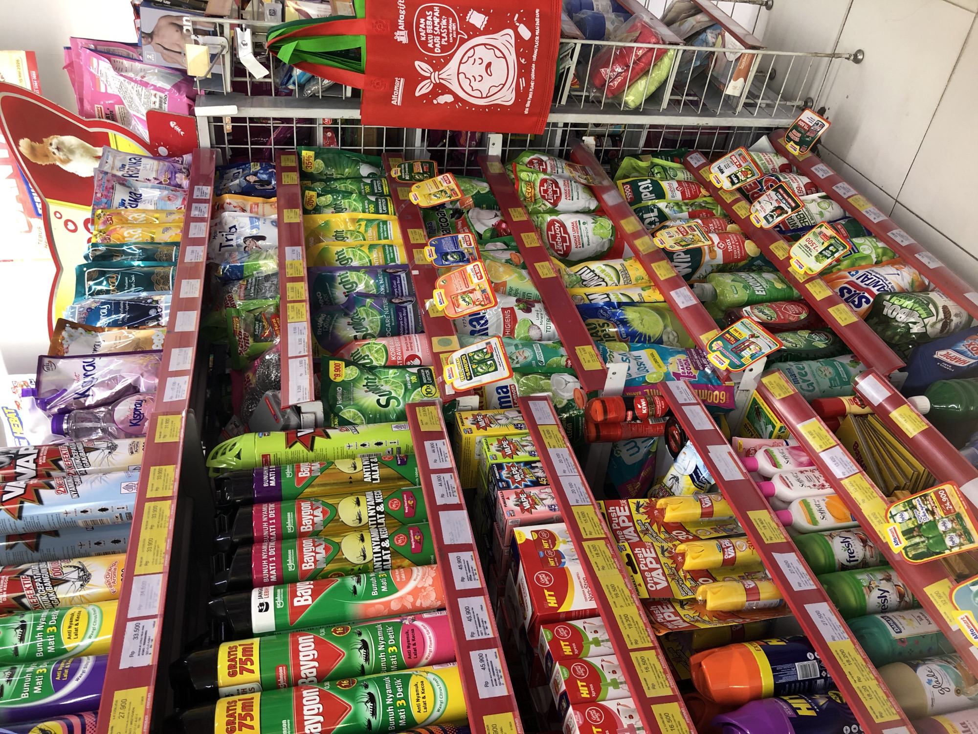 The image shows a retail store aisle in Putat, Indonesia, stocked with various household cleaning and pest control products. The shelves are filled with brands like Baygon, HIT, Kismo, Sunlight, SOS, Dahhila, and VAPE. Products include insecticide sprays, laundry detergents, dish soaps, and air fresheners. Prices are displayed in Indonesian Rupiah (Rp). A red reusable shopping bag with a cartoon drawing of a sad garbage bag and the text "Alfagift" is hanging on the right side of the image. The store appears to be well-lit and organized. There are no people visible in the image.