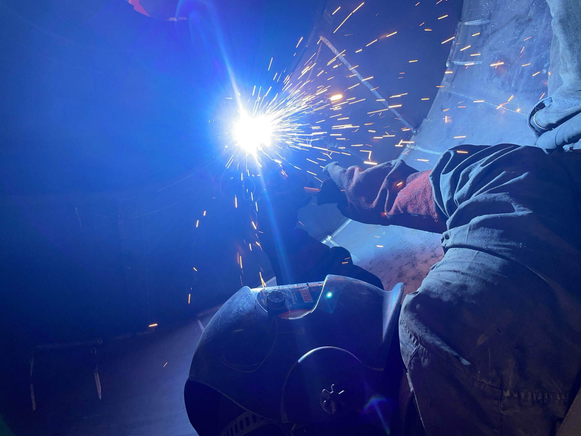 A welder in a protective helmet and suit works on a metal structure, illuminated by the bright arc of welding. Sparks fly from the welding point, creating a dazzling display against the dark, industrial environment. The scene appears to be an interior space, possibly a construction site or workshop, with large, curved metal surfaces visible. The overall impression is one of focused labor in a high-temperature, high-intensity setting. The location in Tomba, Montenegro is not discernible from the visual evidence provided in the image.