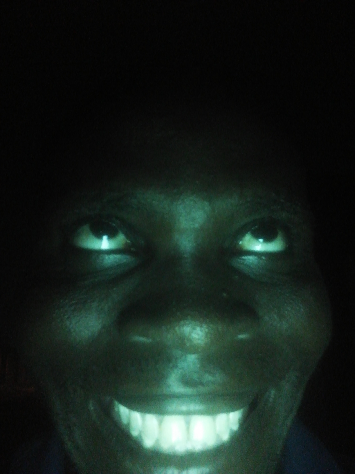 A close-up photograph shows a person's face in low light, seemingly indoors. The person has dark skin, eyes looking upward, and a wide smile revealing illuminated teeth. The background is obscured by darkness, making it difficult to discern further details about the environment.