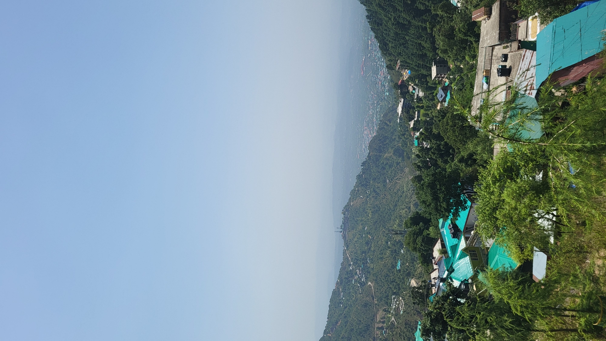 The image shows a landscape in Bhagsu, India. A mountain range with lush green vegetation covers the right side of the frame. Buildings with blue rooftops are nestled amidst the trees. In the distance, a city is visible, shrouded in a light haze. The left side of the frame is dominated by a clear, blue sky.
