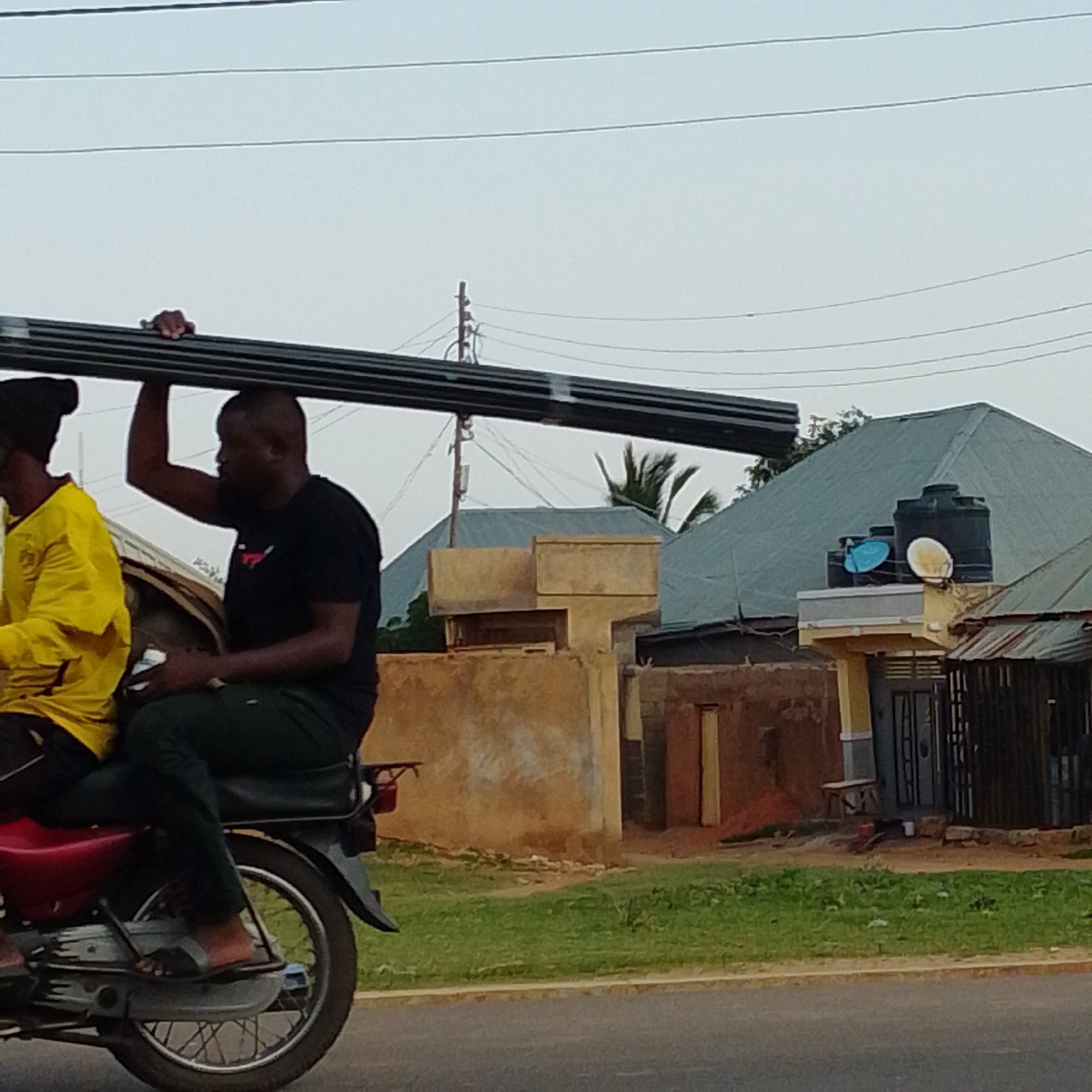 In Gombe, Nigeria, two individuals are riding on a motorcycle, transporting a long, dark object that appears to be metal piping. The person in front is wearing a yellow long-sleeved shirt and a black head covering, while the person behind, driving the motorcycle, is wearing a black t-shirt and dark pants. The motorcycle is red and black. They are traveling on a paved road, with a grassy area and buildings in the background. The buildings have various roofing materials, including what appear to be corrugated metal and asphalt shingles, and some have water tanks on their roofs. The sky is overcast.