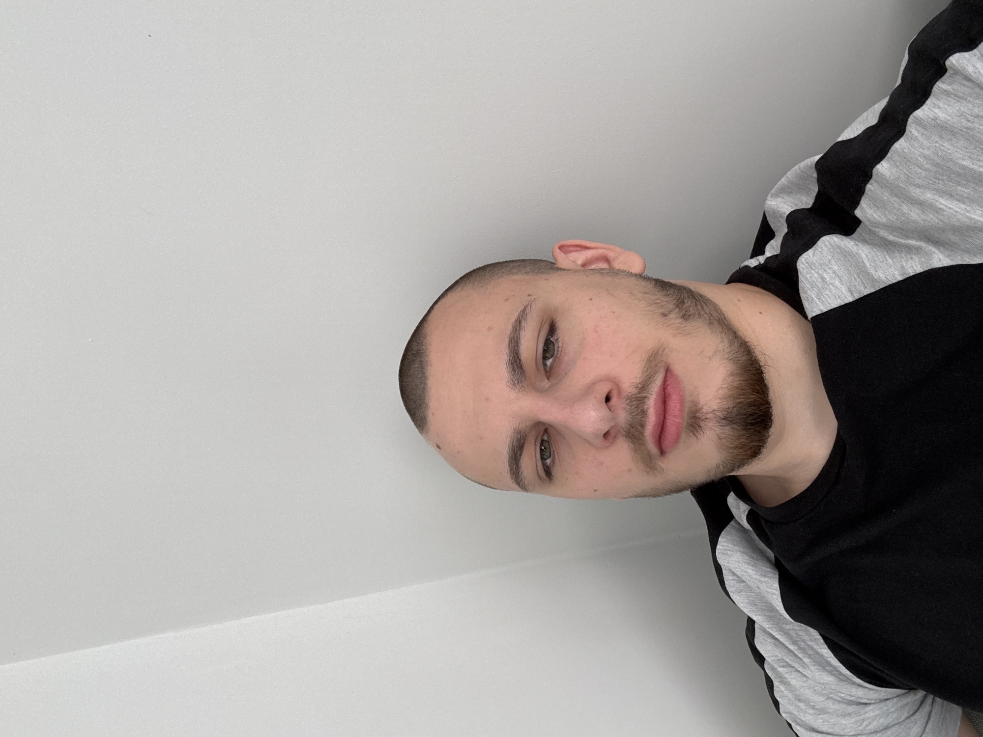 A close-up, angled selfie shows a man indoors. He has short-cropped, dark hair, light skin, and a beard. He is wearing a black and gray long-sleeved shirt. The background is a plain, white wall. The location is unknown and has not been revealed, and there is no information to suggest that it is located in Stara Zagora, Bulgaria.