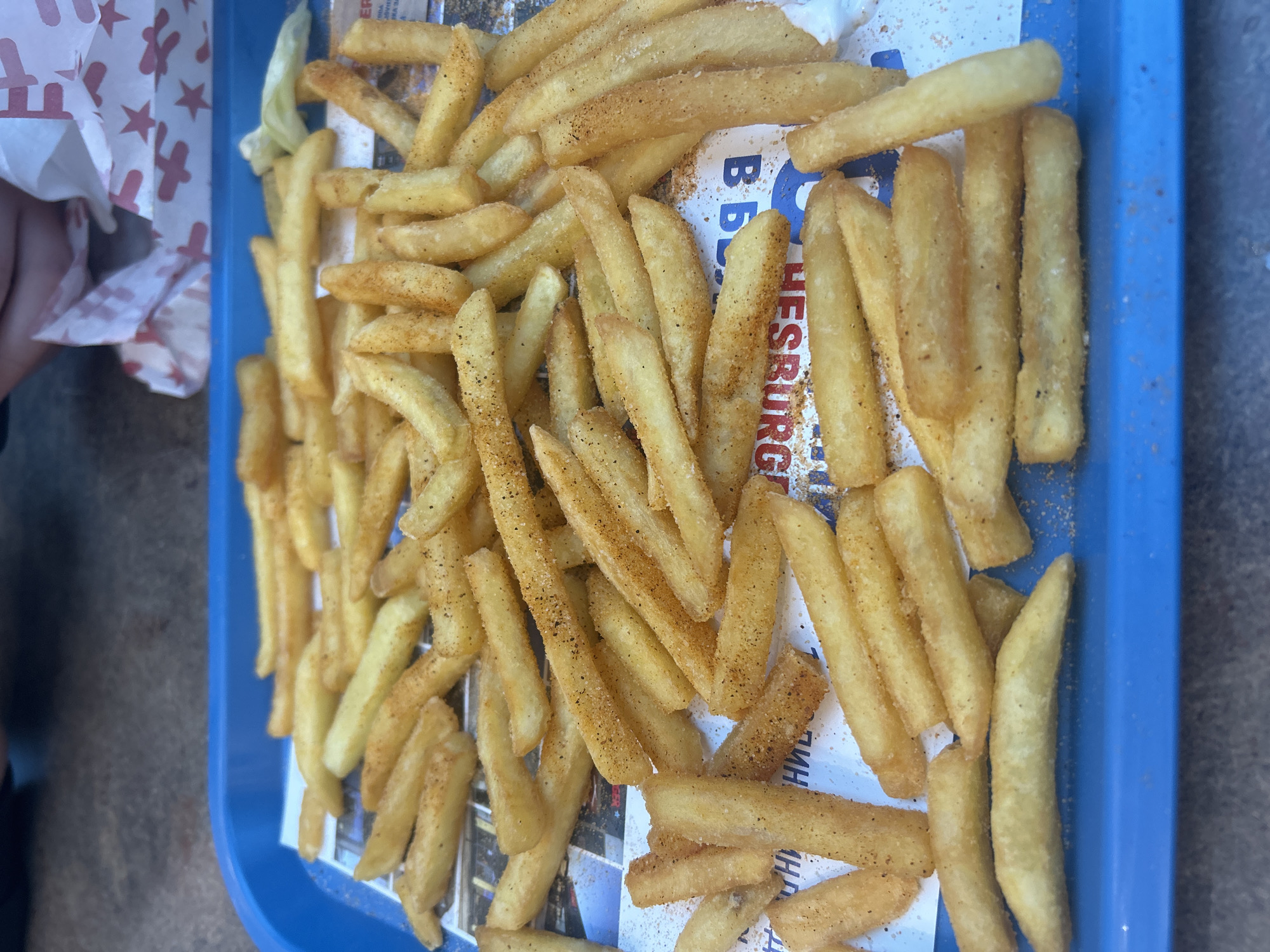 A blue plastic tray is filled with french fries served on a paper liner featuring a logo with the words "HESBURGER", which is a fast-food chain with a location in Varna, Bulgaria. The fries are golden and appear seasoned with a slightly granular, reddish-brown spice mix. To the left, partially visible is a white and red patterned paper bag. The overall image is taken at an angle and looks to be an outdoor shot. The background is a muted grey-beige.
