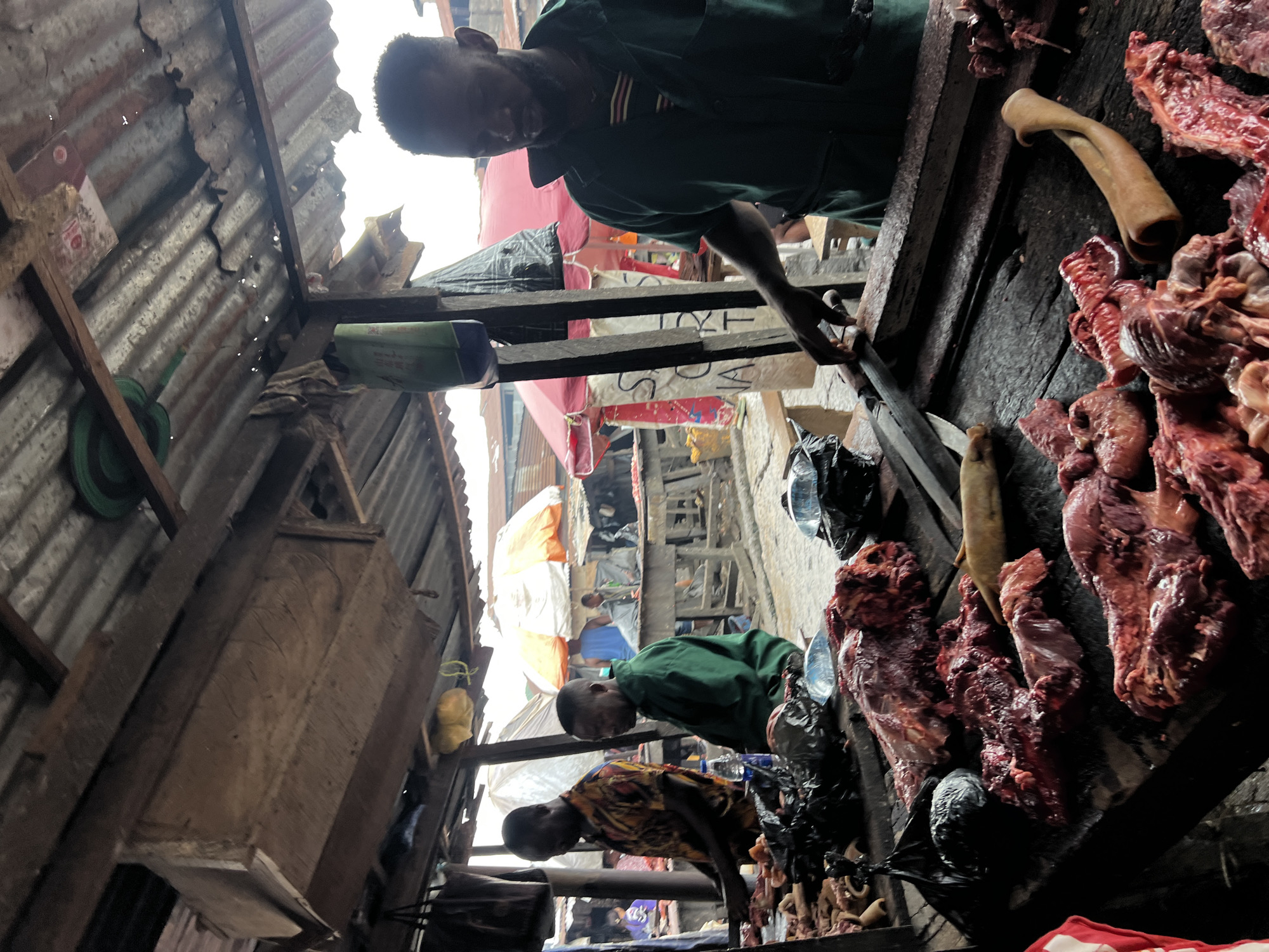 This image depicts a scene at a meat stall in the Nkpor, Nigeria, market. The stall is covered by a corrugated iron roof supported by wooden beams. In the foreground, a butcher, a man with dark skin wearing a dark green jumpsuit, is actively engaged in his work. He is standing behind a wooden counter laden with various cuts of raw meat, including what appears to be beef and pork. The meat is displayed in large portions, some with bone and fat. The butcher is holding a large cleaver and seems to be preparing the meat. To the left of the main butcher, another man in a vibrantly patterned shirt is also attending to meat on the counter. Further in the background, another man in a green shirt is seated. The market environment is visible behind the stall, with other stalls, market-goers, and umbrellas suggesting a busy outdoor marketplace. The overall atmosphere is that of a bustling local food market.