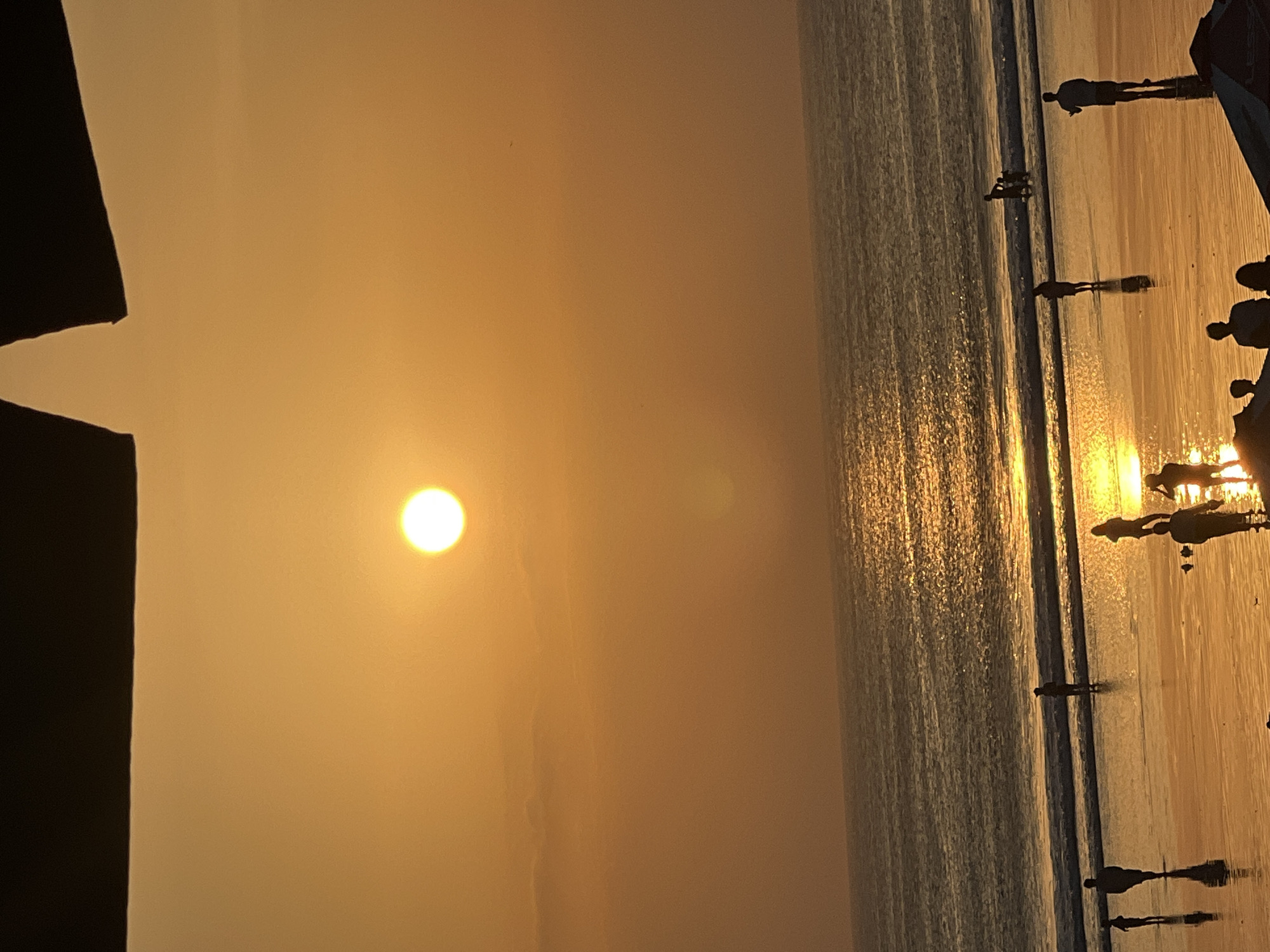 An outdoor, vertical, medium shot captures a beach scene at sunset in Kuta, Indonesia. The sun, a bright, luminous orb, is positioned high in the sky, casting a warm golden glow across the entire frame. The sky transitions from a deep, dark hue at the very top, presumably from an overhead obstruction, to a vibrant orange, gradually softening to a lighter tone towards the horizon. The ocean occupies the middle ground, its surface reflecting the sun's brilliance, with gentle waves lapping at the shore. A wet sand beach stretches across the foreground, also mirroring the sunset's colors and illuminated by the receding light. Silhouettes of several people are scattered across the beach and in the shallow water. Some are standing alone, while others appear to be in small groups, suggesting casual activity and enjoyment of the sunset. The overall atmosphere is serene and tranquil, characteristic of a beach at dusk.