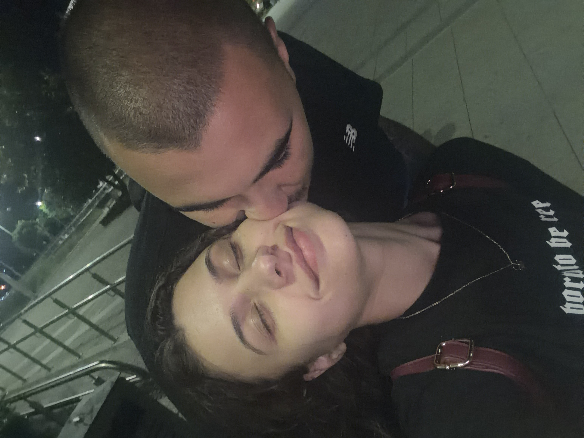 The media file features a close-up, angled shot of two individuals in an affectionate embrace during what appears to be nighttime.
One person, positioned in the upper part of the frame, has a closely shaved head and is wearing a dark hoodie with a white "NB" (New Balance) logo visible on the right chest area. This person is leaning down to tenderly kiss the forehead of the second individual.
The second person, positioned horizontally across the lower part of the frame, has dark, wavy hair and eyes that are gently closed, suggesting a moment of relaxation or contentment. They are wearing a dark long-sleeved top or t-shirt. A silver chain necklace is visible around their neck, and a dark red strap with a silver buckle passes over their right shoulder, possibly from a bag or garment detail. White text in a gothic-style font is partially visible on their shirt, reading "born to be free".
The background is dark and out of focus, indicating a nighttime setting. Blurred lights suggest an urban environment, and metallic rails or bars are faintly visible, hinting at an outdoor location such as a bridge, balcony, or elevated walkway. The overall mood conveyed is one of affection and intimacy between the two subjects.