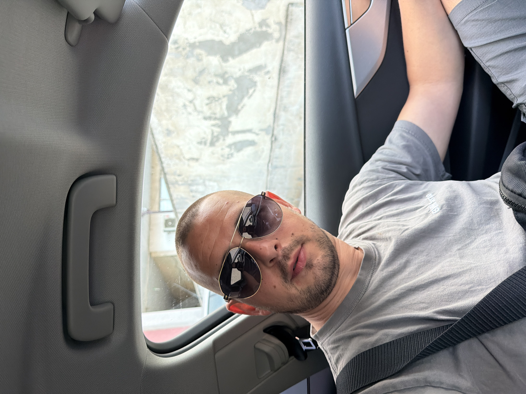A man with a shaved head, wearing sunglasses and a grey t-shirt, is sitting in the passenger seat of a car. He is looking at the camera. The car's interior is visible, including the ceiling handle and the door panel. Through the window, a textured wall and a building can be seen. The image appears to have been taken in Burgas, Bulgaria.