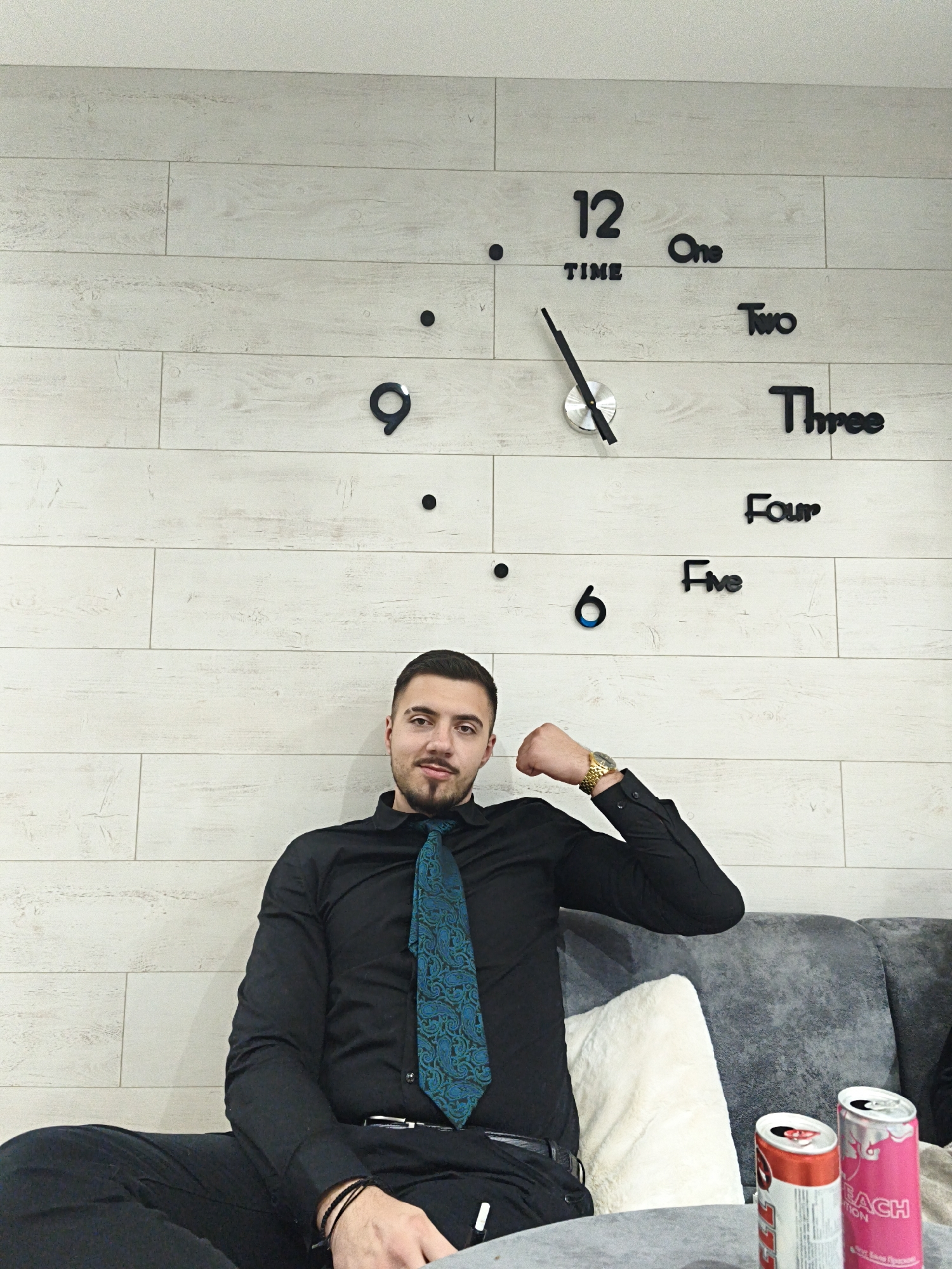 A man is sitting on a couch in front of a light-colored, horizontally-paneled wall with a large, decorative clock. The clock's numbers and hands are black and silver. The man is wearing a black dress shirt with a green and blue patterned tie, and black dress pants. He is wearing a gold watch on his left wrist, which is flexed. There are two opened cans of energy drinks on a table in front of him: one pink "Peach Edition," and one red "Edition". The couch is gray with a white decorative pillow. The setting appears to be indoors. The location, Pleven, Bulgaria, is not identifiable from the image.
