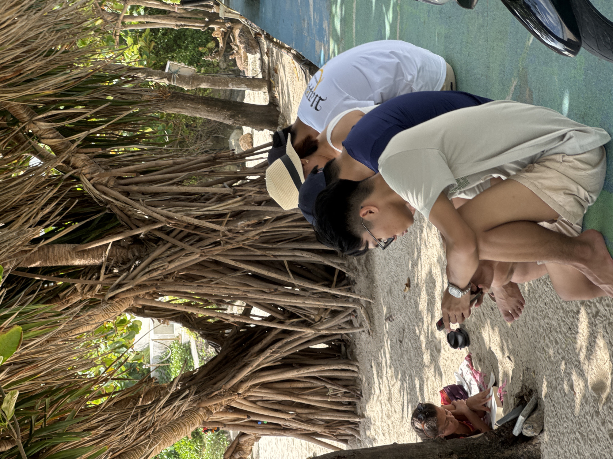 In Patong, Thailand, at a beachside location shaded by a dense canopy of a banyan tree, several individuals are present on a sandy expanse. In the foreground, a person wearing a light-colored t-shirt and beige shorts sits with their legs crossed, looking down at a mobile phone. To their left, another individual in a dark blue shirt is seated, facing away from the camera. Further to the left and slightly behind, a person in a white t-shirt with an intricate graphic on the back is also seated. A person wearing a straw hat with a black band is partially visible between the person in the white t-shirt and the individual in the dark blue shirt. To the far left, nestled against the tree trunk, a person wearing sunglasses and a colorful top is reading a book. Scattered on the sand are a pair of black flip-flops, a small pile of colorful fabric, and some rocks. The background reveals more vegetation and a pathway. A portion of a black motorcycle is visible on the right side of the frame. The overall scene depicts a relaxed atmosphere, with individuals engaged in individual activities under the natural shade.