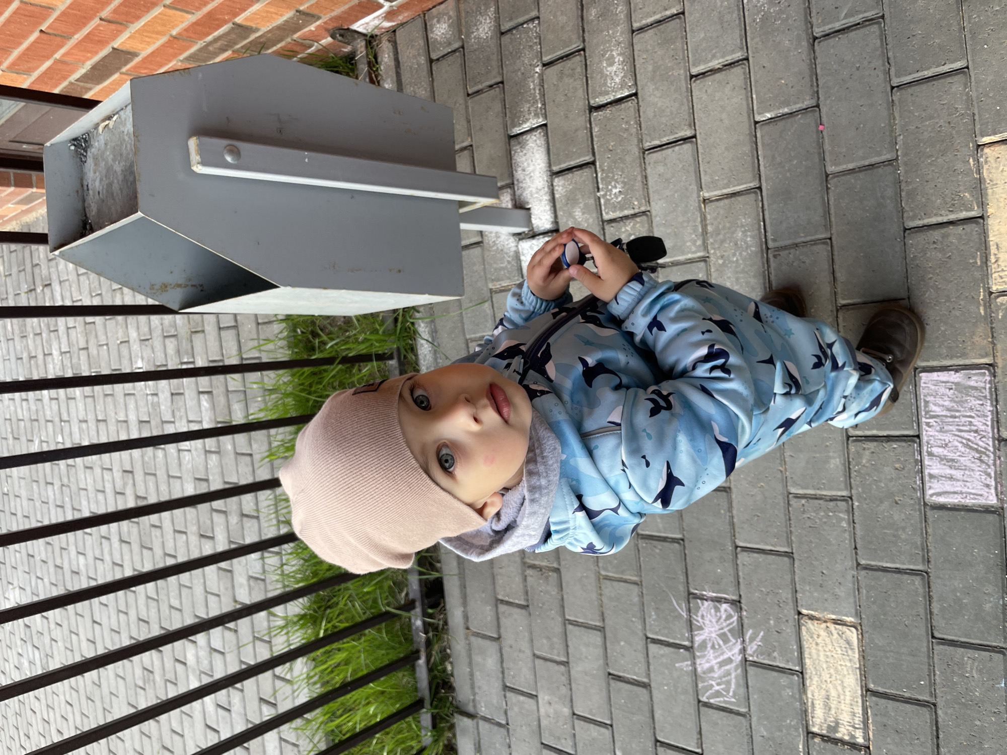 A child in a light blue whale-patterned snowsuit and a beige beanie stands on a gray brick sidewalk. The child holds a small object in their hands. Next to the child is a dark gray metal structure. A low black fence is visible to the left, with green grass growing behind it. In the background, there is a brick building. The location is Izhevsk, Russia.