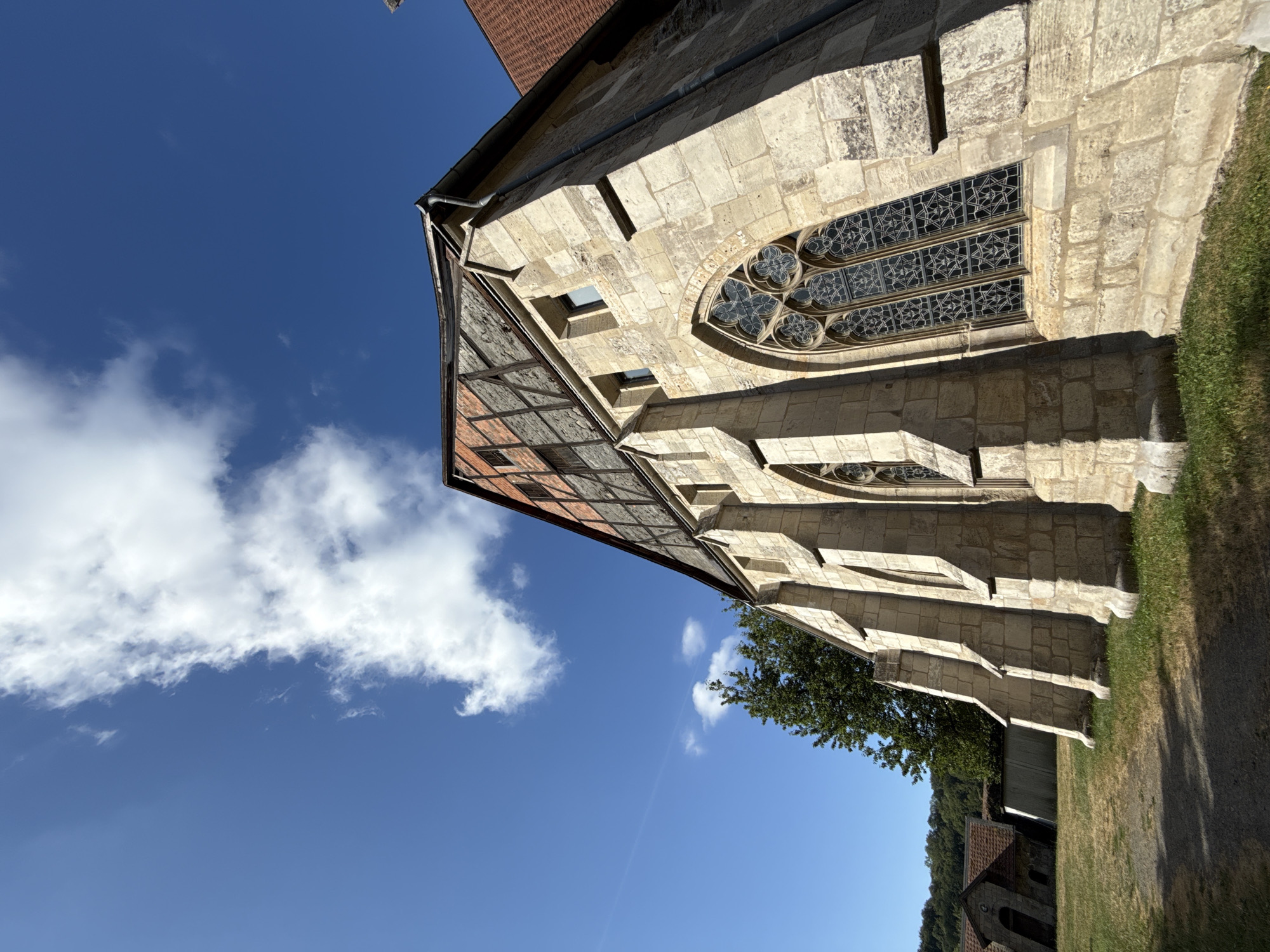 This is a low-angle, wide shot of the exterior of a stone church, likely St. Mary's Church in Walkenried, Germany, on a sunny day. The architecture is Gothic, with pointed arches and buttresses visible on the side of the building. A large, ornate Gothic window with dark panes and intricate stonework is prominent. The upper part of the building features a timber-framed section with exposed wooden beams and brick infill, topped by a reddish-brown tiled roof.
The church is situated on a grassy area with patches of dry, straw-like grass. A dirt path runs in the foreground, casting a shadow from the church. To the left of the church, in the background, are other older buildings with similar tiled roofs, one appearing to be an arched entrance. A dense green tree stands behind the church, and a wooded hillside is visible in the far distance. The sky is a vibrant blue with a few scattered white clouds. No people are present in the image.