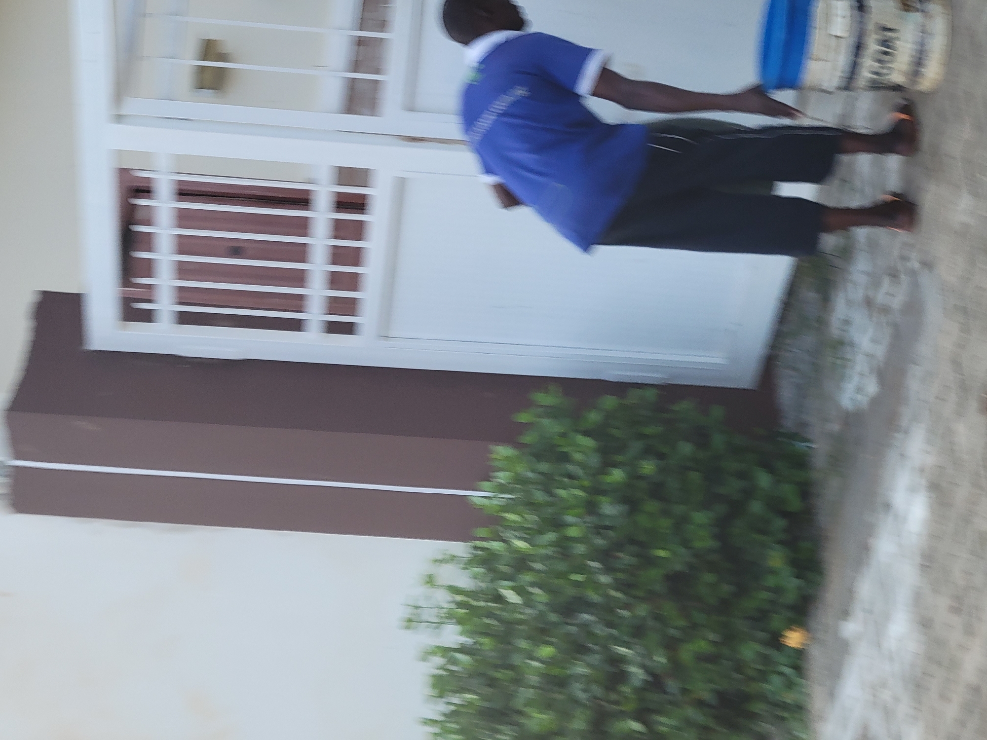 The image depicts a male individual, appearing to be of African descent, wearing a blue t-shirt with white trim on the collar and sleeves, and dark-colored, loose-fitting trousers. He is also wearing open-toed sandals. The individual is bent over, with his right hand near a large white bucket with a blue lid. The bucket has some text on it, including what appears to be "GOAT". The scene is set outdoors, in front of a building with a white wall. To the left of the building is a large green bush. Adjacent to the building is a white metal gate with vertical bars. A brown, rectangular architectural element is also visible, extending from the top of the frame downwards along the left edge of the gate. The ground appears to be a mix of dirt and possibly some gravel. The lighting suggests it is daytime. The setting is consistent with an outdoor area in Abuja, Nigeria, possibly a residential or commercial property undergoing some form of work or maintenance.