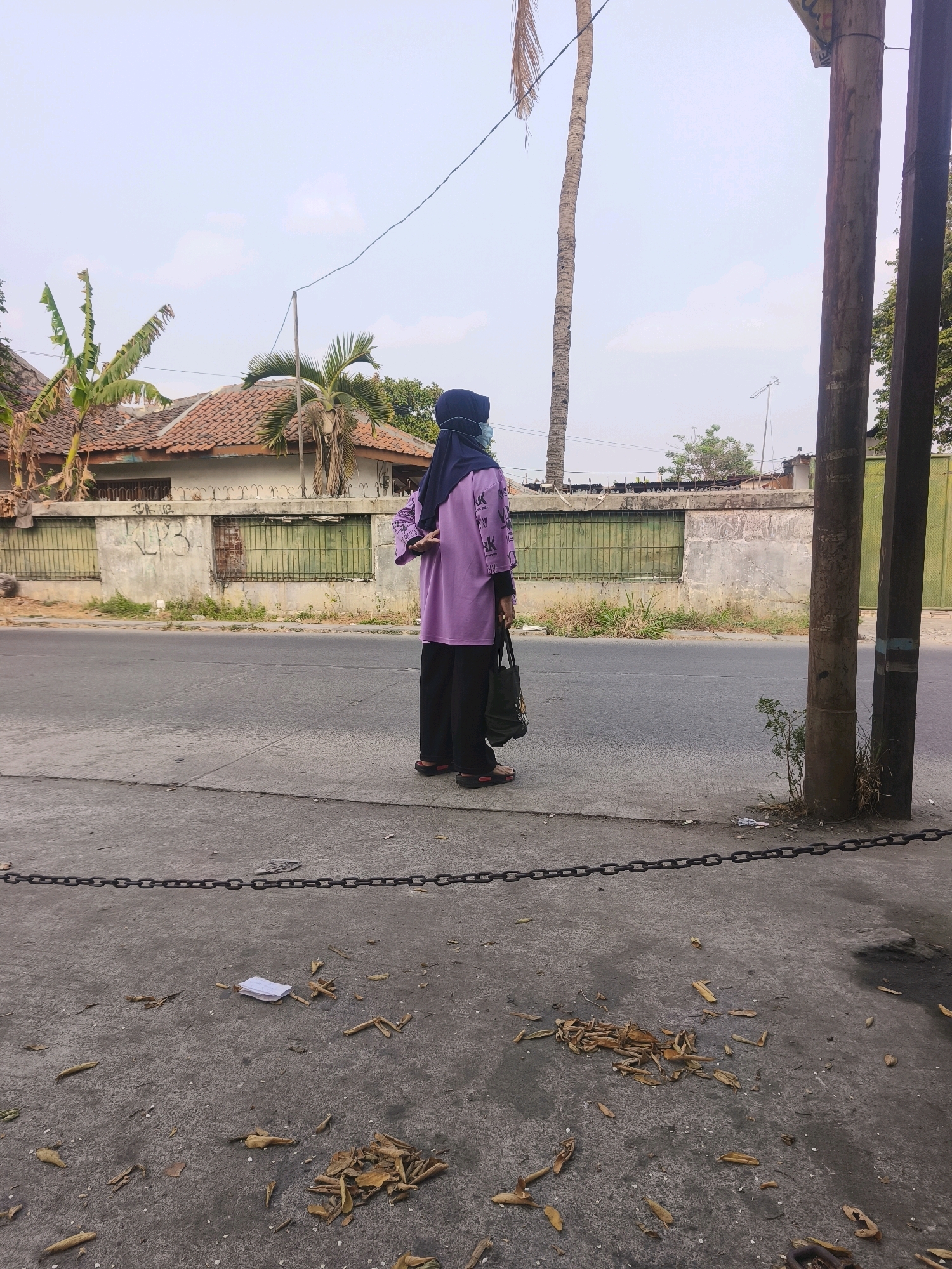 A person wearing a purple hijab and a long-sleeved purple shirt with black lettering and black pants stands on a sidewalk facing away from the camera. The person is also wearing a light blue face mask and sandals. They are holding a dark-colored tote bag. In the background, there is a paved road, a low wall with graffiti, green metal gates, a house with a tiled roof, banana trees, and palm trees. There are also power poles and wires in the scene. The ground in the foreground is concrete and is littered with dry leaves and a piece of white paper. The scene appears to be in an outdoor urban or semi-urban environment, likely in Bekasi, Indonesia, as suggested by the presence of tropical vegetation and architectural styles.