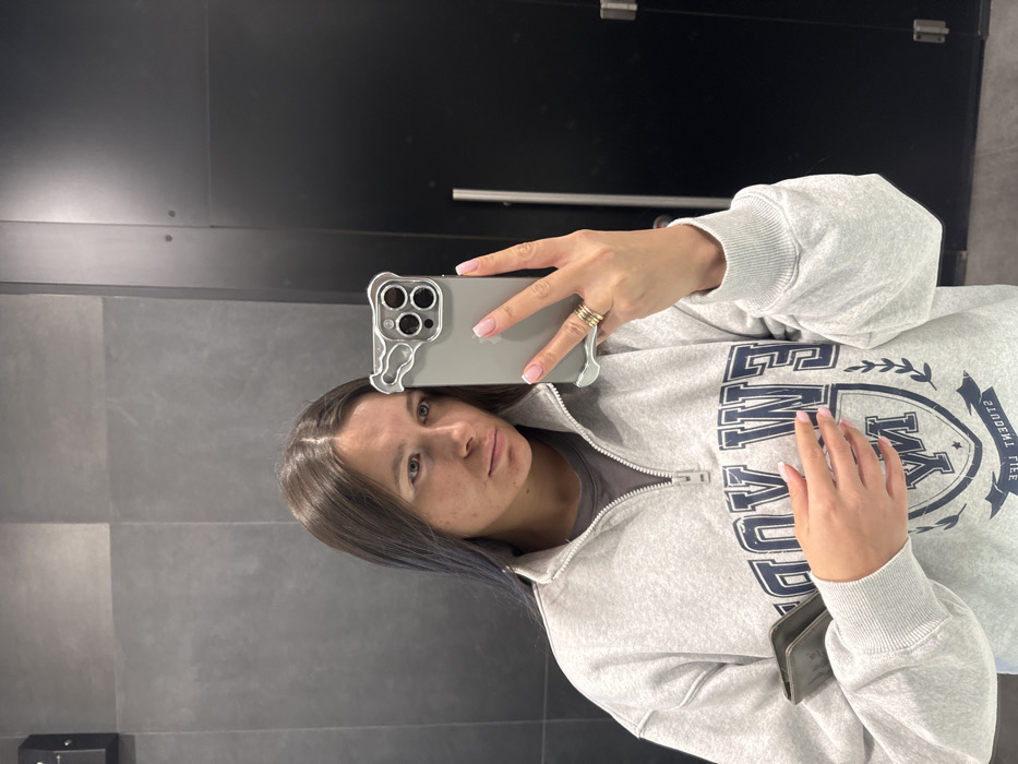 An indoor mirror selfie shows a young woman looking directly at the camera. The image is taken at a 90-degree angle, with the subject's head tilted to the right. She has dark, straight hair, parted down the middle, with a subtle blue tint visible in some strands near her face. Her expression is neutral, and her skin shows a few freckles or minor blemishes.
She is wearing a light grey, long-sleeved sweatshirt with a quarter-zip collar, revealing a darker grey or black t-shirt underneath. The sweatshirt features a prominent dark blue graphic on the front: a crest or shield design with a large "M" in the center. Above the "M," the word "MIAMI" is visible, and below it, "WE." Along the right side of the crest, oriented vertically, the words "STUDENT LIFE" are printed.
The woman is holding a smartphone, likely an iPhone, with her right hand, which is adorned with several rings on one finger. The phone has a distinctive clear or silver-toned case with wavy edges around the camera module and along the sides. Her fingernails have a classic French manicure with light pink polish and white tips. Her left hand is resting casually on her stomach, also showing the French manicure.
The background consists of dark, possibly charcoal grey, tiled or paneled walls with horizontal seams. In the upper portion of the background, a very dark, almost black, shiny surface (possibly a door or another wall section) is visible. A small, dark rectangular object is present on the lower left wall, possibly a dispenser or sensor. The lighting appears even and artificial, suggesting an indoor setting like a restroom or hallway.