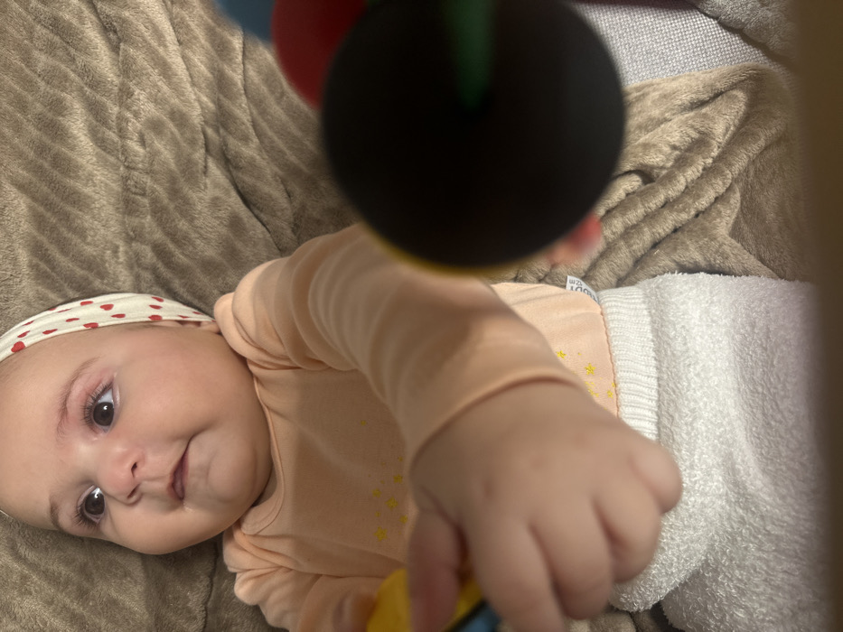 A baby wearing a headband with red hearts is lying on a soft, brown blanket. The baby is wearing a light orange shirt with yellow star patterns. A hand reaches towards a black, red, and green toy. The baby is also wearing white bottoms. The baby is looking at the camera.