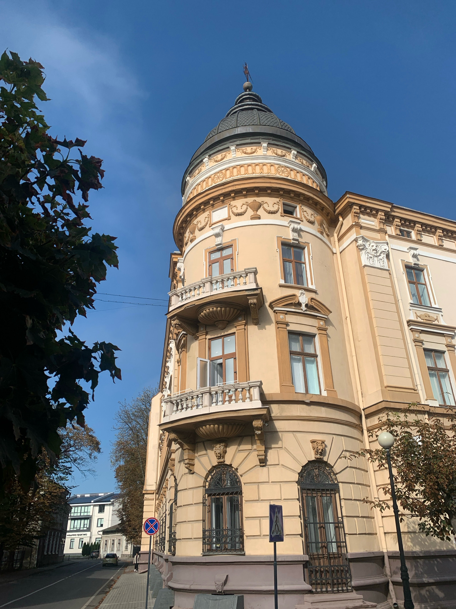 The image captures a warm, sunny day in Kolomyia, Ukraine, focusing on a building with distinct architectural features. The building, painted in a pale yellow hue, is adorned with ornate detailing, including a rounded turret topped with a dark dome. Balconies with white railings punctuate the facade, and arched windows with decorative wrought-iron grilles are visible on the lower levels.
To the left of the building, a leafy tree with some reddish-brown foliage casts a shadow, suggesting it might be autumn. A paved sidewalk runs alongside the building, and a street stretches into the distance, lined with more buildings and trees. A car is parked on the street, and a few signs are present near the sidewalk, including a blue sign with a white diagonal line and a red circular sign with a blue cross. A lamppost with a spherical light fixture stands near the corner of the building. In the background, another modern building with a white facade and dark-framed windows is visible. The sky is a clear, vibrant blue. The overall scene depicts a street view in a city, with an emphasis on historic architecture.