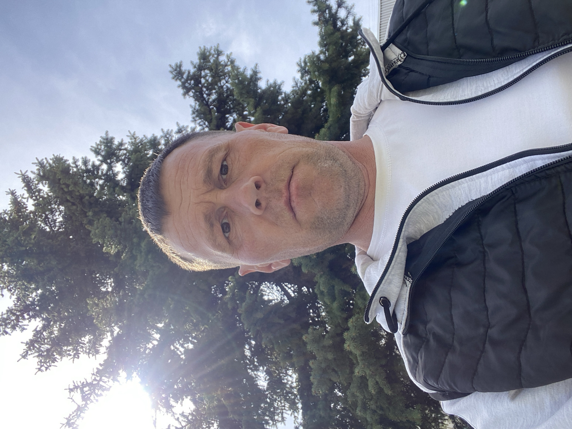 A selfie taken by a man with short hair, wearing a white t-shirt, a grey zip-up hoodie, and a black quilted vest. The background shows evergreen trees under a partly cloudy sky with the sun shining through the branches. The location is not discernible as Chaikovsky, Russia.