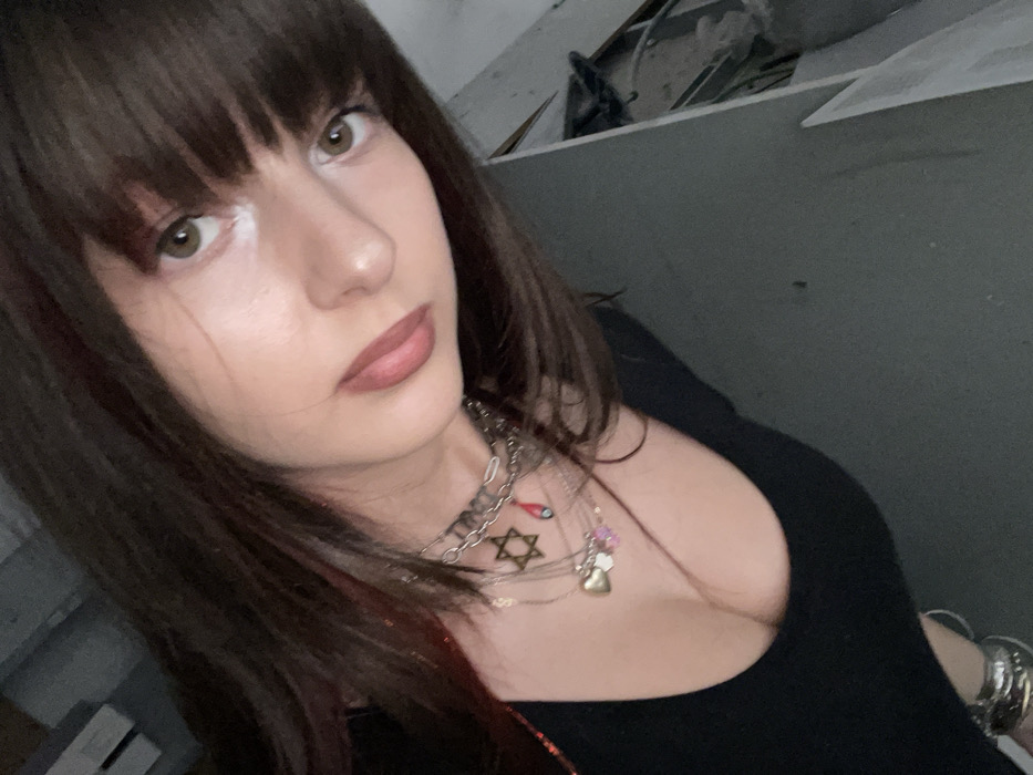 A young female individual is depicted in a medium close-up shot, angled slightly from above, looking directly at the camera. She has long, dark brown hair with straight bangs covering her forehead and reddish undertones visible in some strands. Her eyes are light brown, and she has white cosmetic liner under the inner corner of her right eye. Her lips are a muted pinkish-brown. She wears a black, low-cut top or dress. Layered necklaces are visible around her neck: a metallic choker, a chain with a Star of David pendant, another chain with a red teardrop-shaped pendant and a small blue charm, and a longer chain featuring a heart-shaped locket with smaller, light-colored charms. Several silver-toned bracelets adorn her left wrist. The background is dark and appears to be an interior space, with a plain dark gray wall behind the subject and indistinct, possibly cluttered or damaged architectural elements to her right. The scene is located in Raanana, Israel.