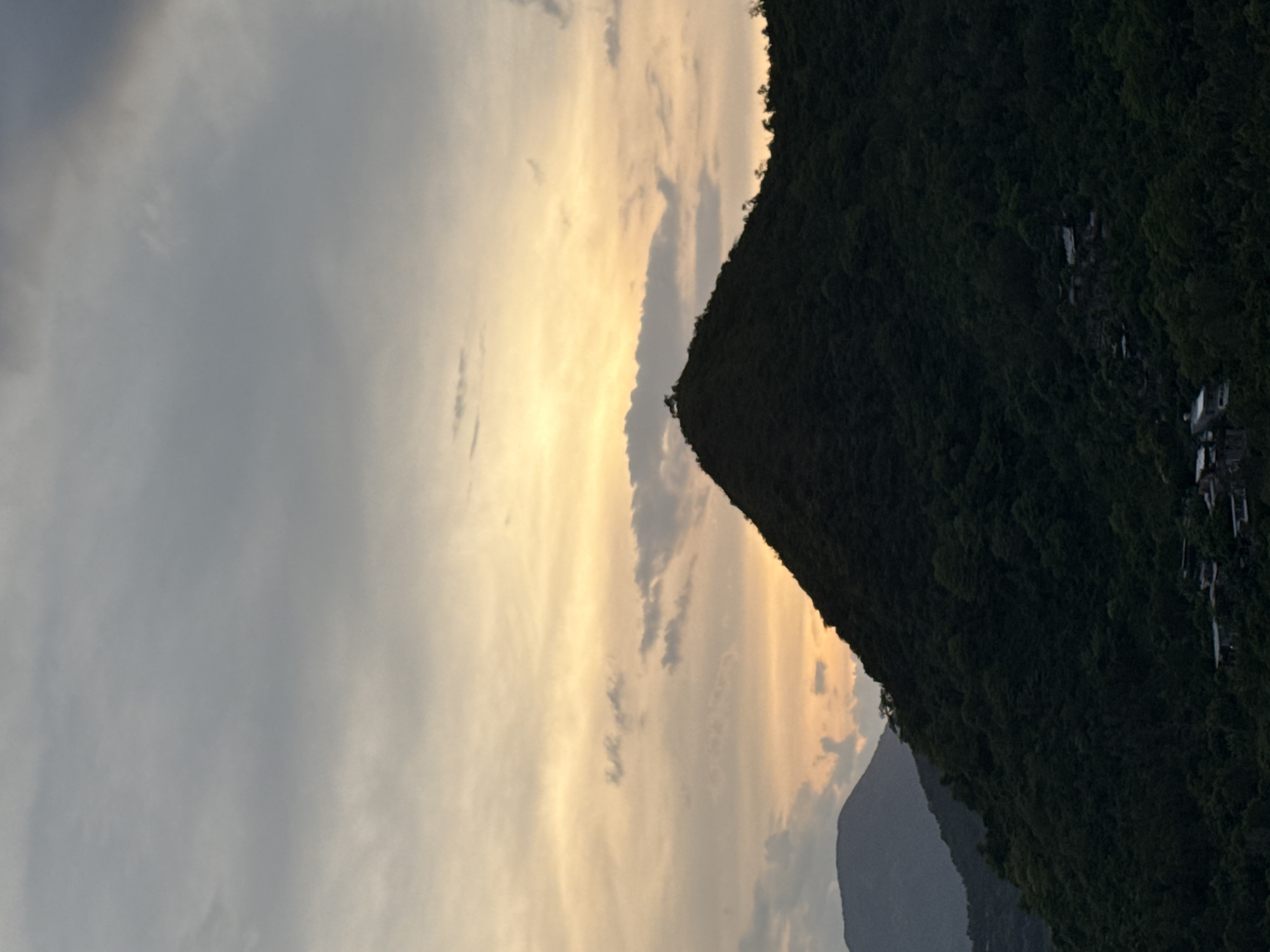 The image shows a mountainous landscape at dusk in Bac Natural Heritage Area, Trang, Vietnam. The scene is dominated by a steep, densely forested mountain covered in dark green vegetation that stretches across the right side of the frame. The peak of this mountain is silhouetted against a sky filled with clouds.
The sky on the left side of the image displays a gradient of colors from grey near the top to pale yellow and orange as it meets the mountain's silhouette. The soft, diffused light suggests the sun is either setting or has recently set, casting a warm glow. Scattered clouds are visible, some tinged with the sunset colors, while others appear grey and overcast.
In the distance, another, less distinct mountain range is visible, appearing as a muted grey silhouette. Small structures, possibly houses or buildings, are scattered along the upper slopes of the main mountain on the right, adding a touch of human presence to the natural setting. The overall mood is serene and tranquil, capturing the quiet beauty of the natural landscape as day transitions into night. No text is visible in the image.