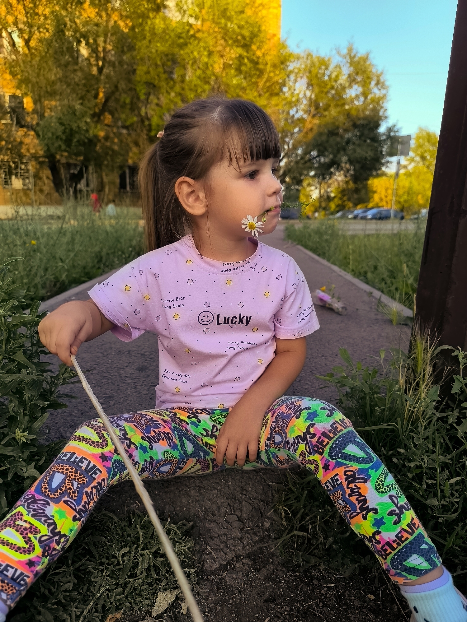 A young girl with dark hair pulled into a ponytail and bangs is sitting on a paved path outdoors. She is wearing a light pink t-shirt with a pattern of small stars and a smiley face, and brightly colored leggings with a "Believe" slogan. A daisy is held between her teeth, and another is partially visible near her mouth. She is holding a thin, light-colored stick in her right hand. The background shows trees with yellow and green leaves, suggesting autumn, and a building. The scene appears to be in a park or urban green space.