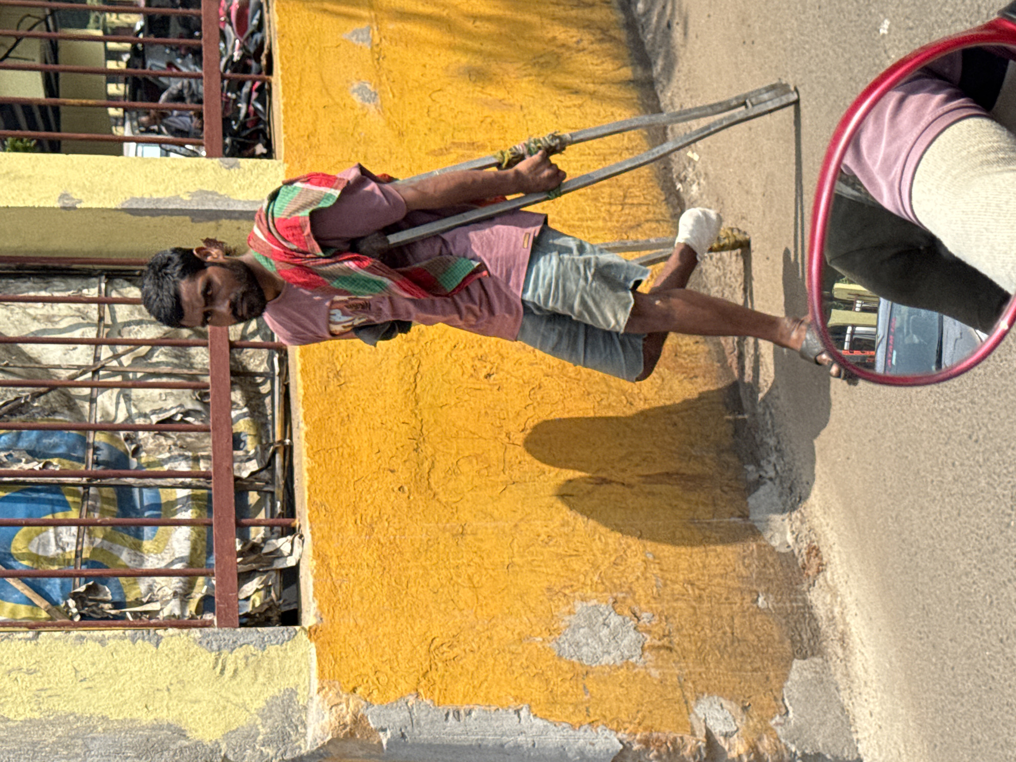 A man stands on a street in Kolkata, India, using crutches. He is wearing a reddish-purple shirt, denim shorts, and a striped shawl. One of his feet is bandaged. He stands next to a yellow wall with a barred window. A red mirror reflects the man's arm and the street scene behind him. The ground is asphalt.
