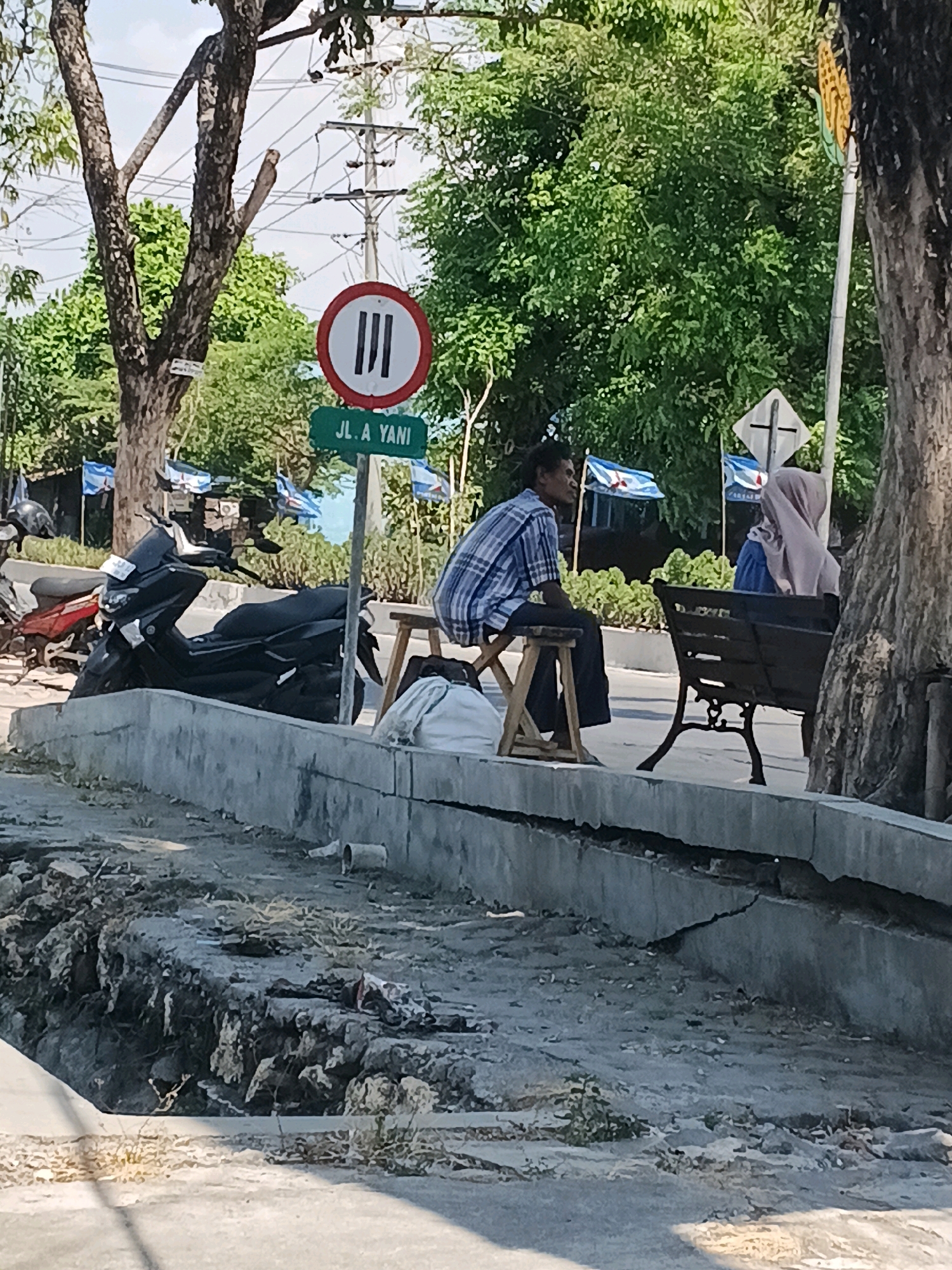 The image depicts a street scene in an urban environment, possibly in Indonesia given the street sign "JL. A YANI" and the flags. The foreground shows a damaged concrete curb and a ditch filled with debris, including plant matter and some discarded items like a plastic pipe and fabric. Beyond the curb, a man in a checkered shirt and dark pants sits on a small wooden stool. To his right, a woman wearing a pink hijab and blue top sits on a park bench. Behind them, several flags with blue and white patterns are visible, along with a traffic sign indicating three vertical lines within a red circle, suggesting a prohibition. Motorcycles are parked on the left side of the scene. Trees and electrical poles are present in the background, with lush green foliage suggesting a warm climate. The lighting indicates it is daytime, likely with bright sunlight.