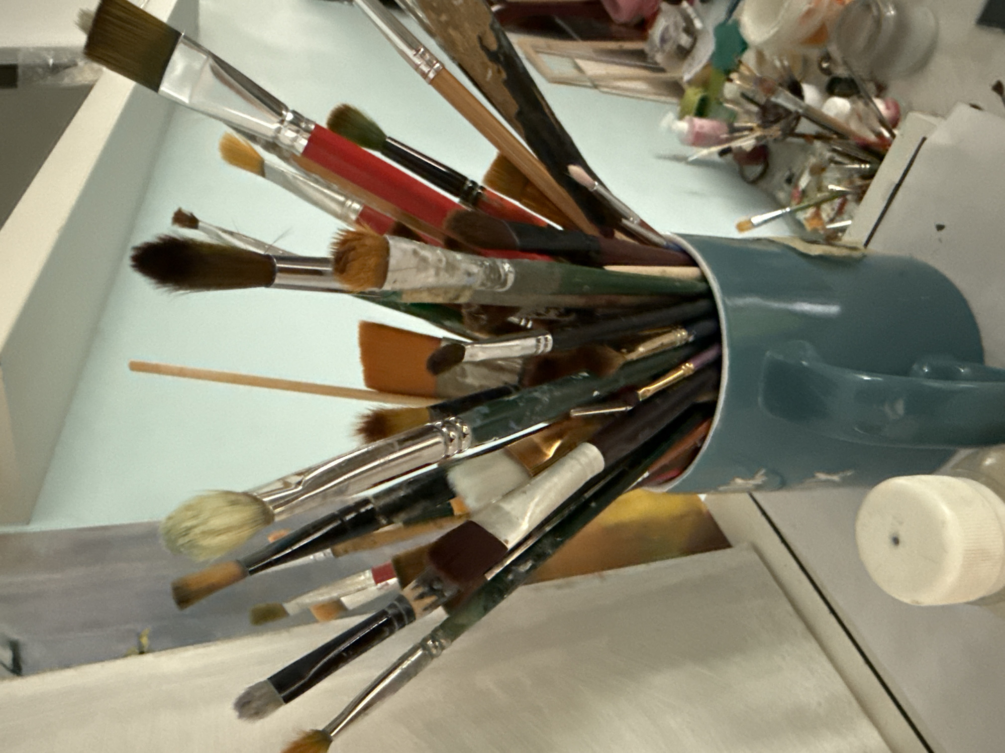 This is a close-up, low-angle photograph of a collection of paintbrushes stored in a teal ceramic mug. The brushes are varied in size and bristle type, with some appearing to be natural hair and others synthetic. Many of the brushes show signs of use, with paint residue on the bristles and handles. The mug is full, with brushes extending out at various angles.
In the background, out of focus, is a table or surface with additional art supplies. There are several small jars and containers, some appearing to hold pigments or mediums. A white cap from a bottle is visible in the lower right of the frame. The overall impression is of an artist's workspace, likely a studio, given the array of materials. The lighting suggests it might be daytime, possibly indoors with natural light. There is no visible text in the image. The image appears to be taken in Vladivostok, Russia.