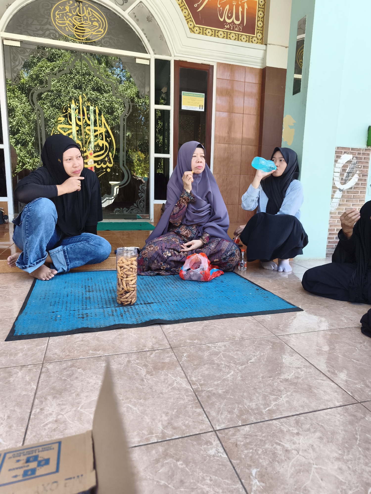 In Tonasa, Indonesia, four women are gathered outside a building, possibly a mosque, given the Arabic calligraphy on the glass doors. Three women are visible in the foreground, sitting on a blue mat on a tiled floor. One woman, wearing a black hijab and jeans, is seated on the left, looking to her right with her hand on her chin. Next to her, a woman in a purple hijab and patterned dress is seated, looking down and holding a snack. A third woman, in a black hijab and a light blue top, is crouching and drinking from a blue water bottle. A fourth woman, also in a black hijab, is partially visible on the far right, her hands clasped in front of her. A jar of snacks and a plastic bag of red items are placed on the mat. The environment appears to be an outdoor, shaded area with a tiled floor and a building with brown wooden doors and pastel blue walls behind them. Sunlight casts shadows on the floor.