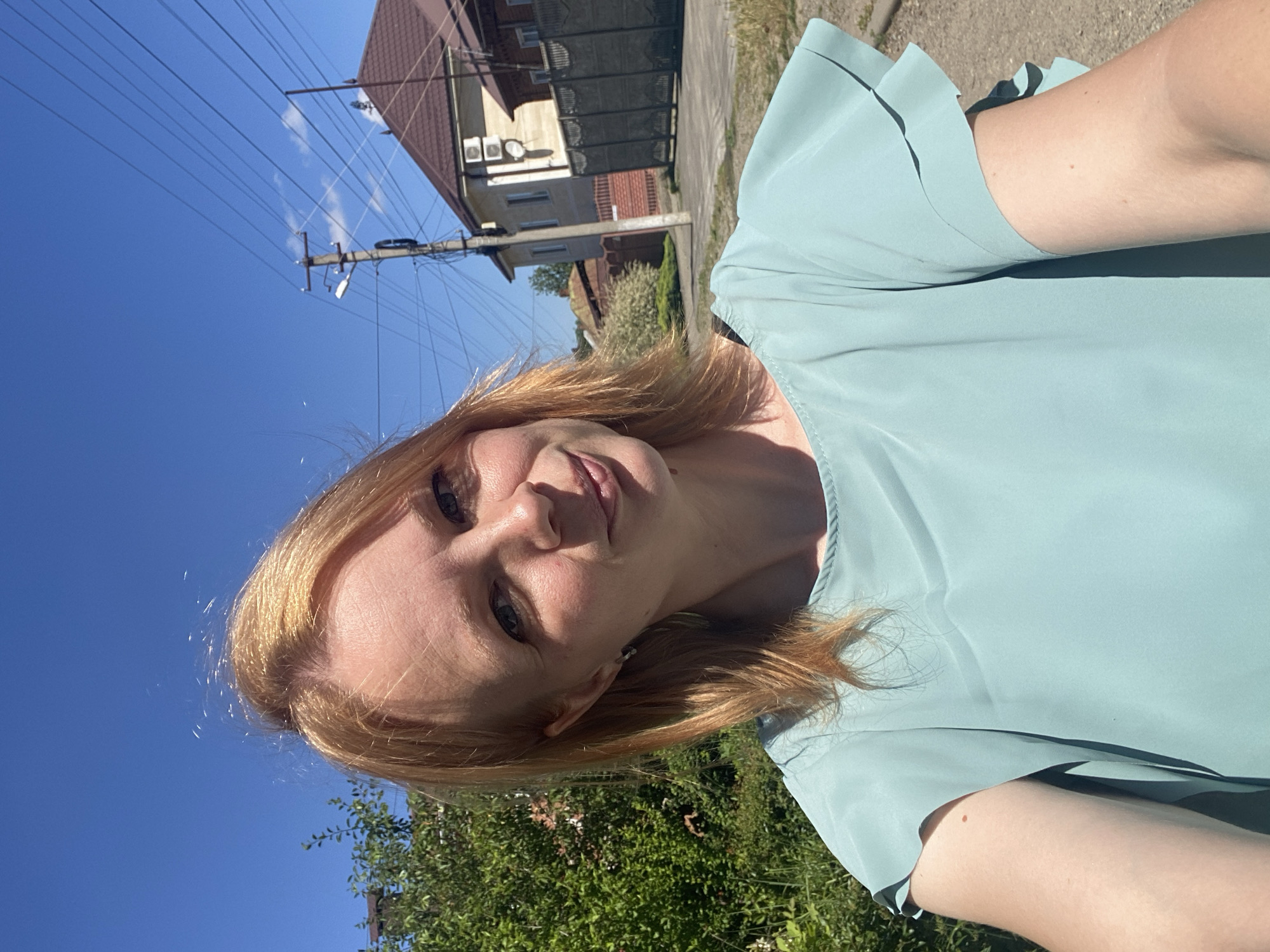 A fair-skinned woman with light red hair is taking a selfie outdoors. She is wearing a light blue-green ruffled top. The background shows a bright blue sky, green foliage, a telephone pole with wires, and houses with brown roofs. A dirt road is visible on the right side of the image. The location is the city of Zapadny, Russia.