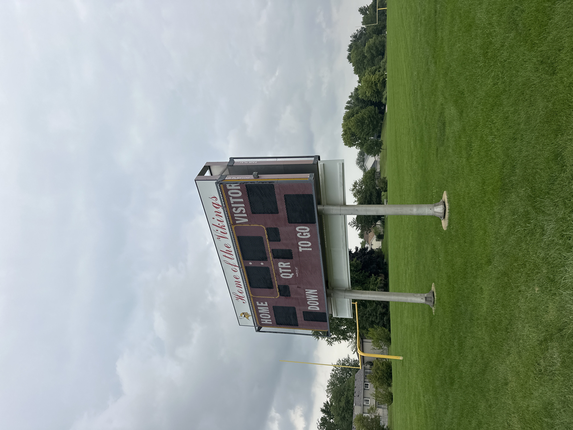 An outdoor, eye-level, wide shot captures a scoreboard at a green, grassy sports field under a cloudy, overcast sky. The scoreboard is positioned in the center of the frame and is supported by two silver poles. The scoreboard itself is large and maroon with yellow trim. At the top, in red script, it reads "Home of the Vikings" with a small Viking mascot graphic to the left. Below this, the scoreboard is divided into sections labeled "HOME," "VISITOR," "DOWN," "QTR," and "TO GO." The score and quarter are not visible as the displays are blank and dark. The field is a vibrant green, with some trees and houses visible in the background. A yellow goalpost is visible to the left of the scoreboard. The overall scene suggests a football field, possibly in a park or school setting. The location is identified as Streamwood, United States of America.