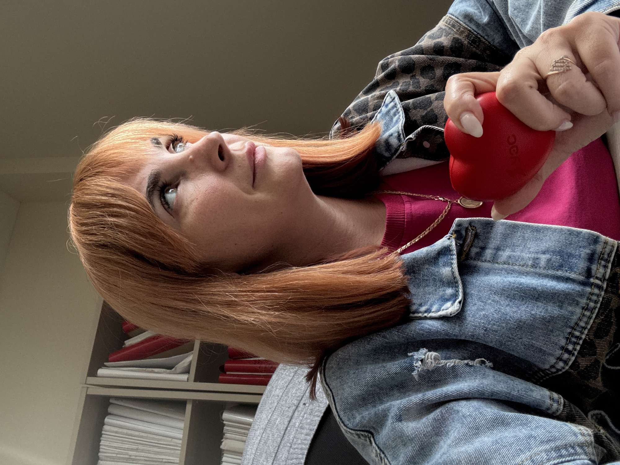 A woman with reddish-orange hair is shown from the chest up, holding a red heart-shaped stress ball in her right hand. She is wearing a bright pink shirt and a denim jacket with a leopard print lining. A gold chain necklace is visible around her neck. Her expression is pensive as she looks upwards and to the left. The background consists of a bookshelf filled with papers and what appear to be red folders or binders. The lighting suggests it is daytime, possibly indoors given the shelving unit. There is no clear indication of the weather. The visible text on the stress ball is "OCD". The image appears to be a self-portrait taken indoors.