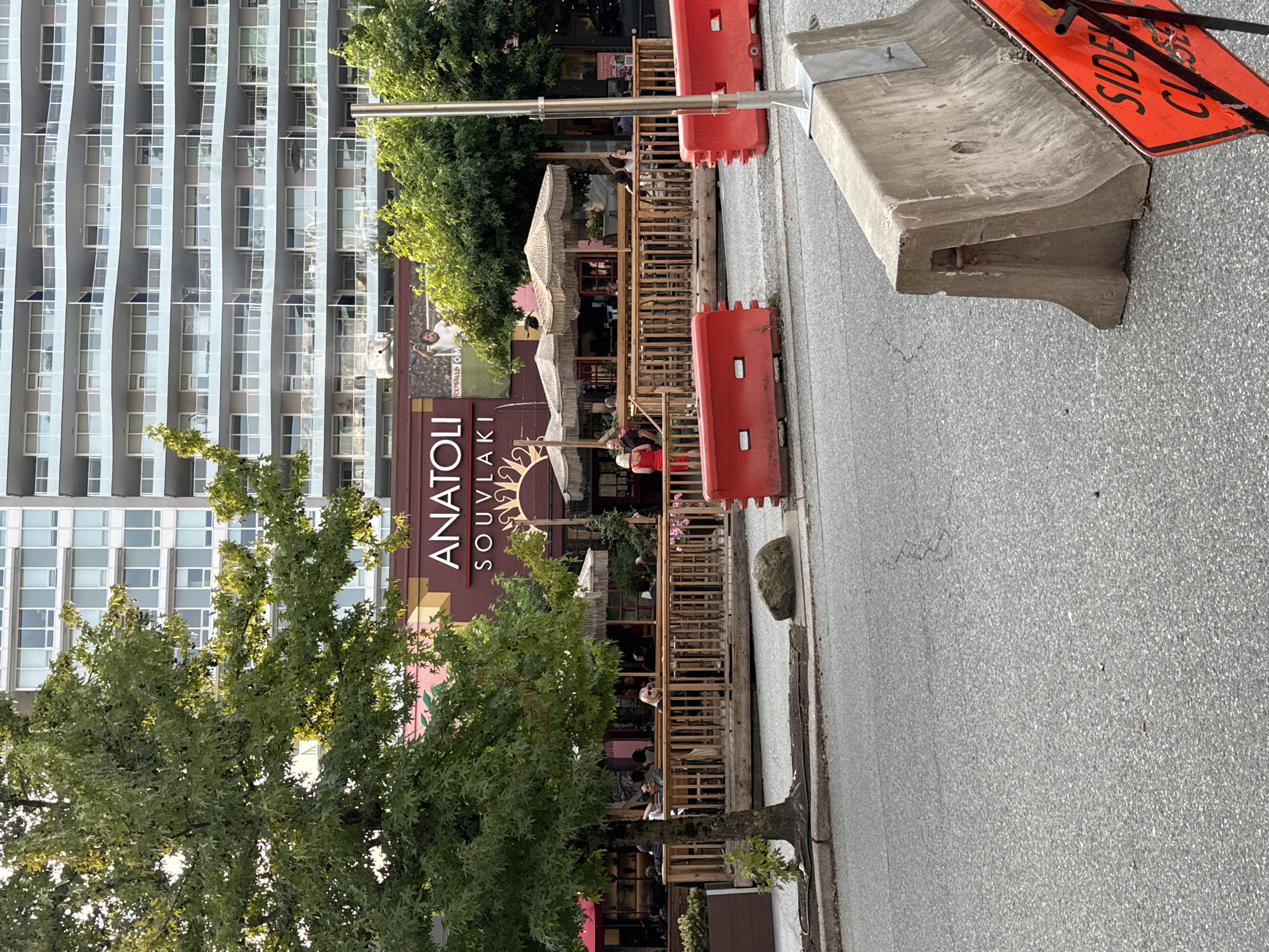 The image shows an outdoor dining area for a restaurant called "ANATOLI SOUVLAKI" in North Vancouver, Canada. A large, modern apartment building with many balconies serves as the backdrop. The dining area is set up on a wooden deck with railings, furnished with tables and chairs under thatched umbrellas. Several people are seated at the tables, appearing to be dining or socializing. In the foreground, there are orange traffic barriers and a concrete barrier with a "SIDE CLOSED" sign, suggesting ongoing construction or road work. A tree with green leaves is positioned to the left of the dining area, partially obscuring the view of the building. The overall scene depicts a lively urban setting with a restaurant operating outdoors.